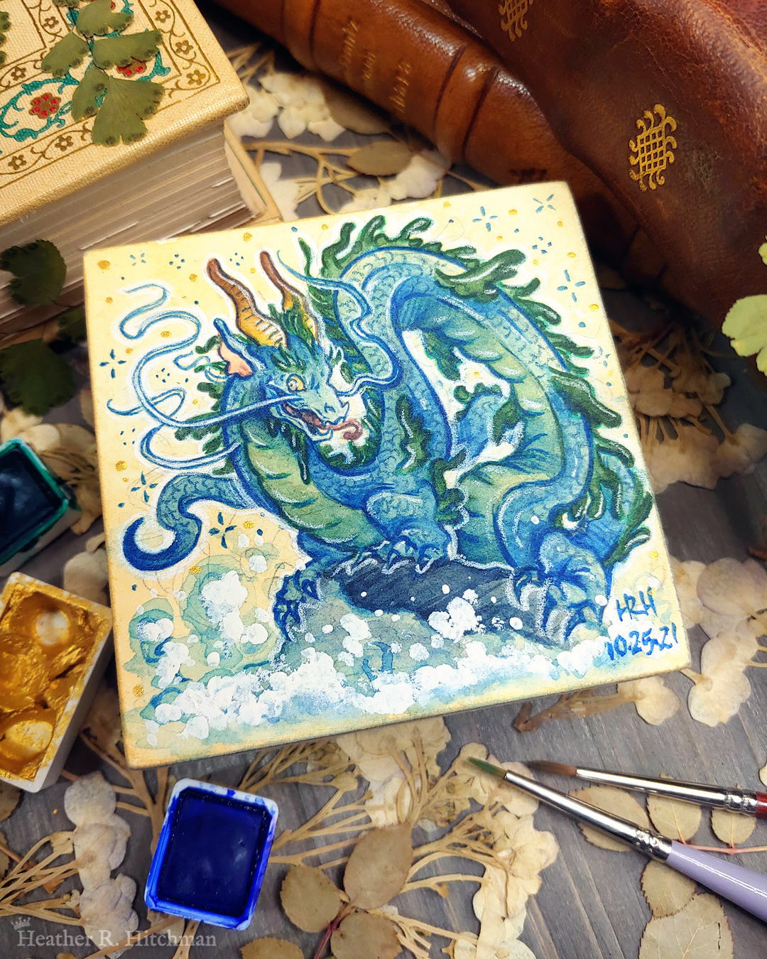 Photograph of a watercolor and gouache painting of an evil rotten leviathan blue water dragon. The dragon has a long, snake like blue and green body with no wings. His body coils across the 3x3 paper against a soft sepia background filled with small gold and blue stars. He is standing on a rock with waves crashing up against it. The dragon’s face has 2 long whiskers that float up from his cheeks to above his head. He has golden goat like horns that twist up into the air. The dragon’s torso is facing the viewer, his face is in profile as he looks towards the viewer with a menacing grin. The painting sits on top of a table covered with watercolor paint pans, books, and foliage in the background.