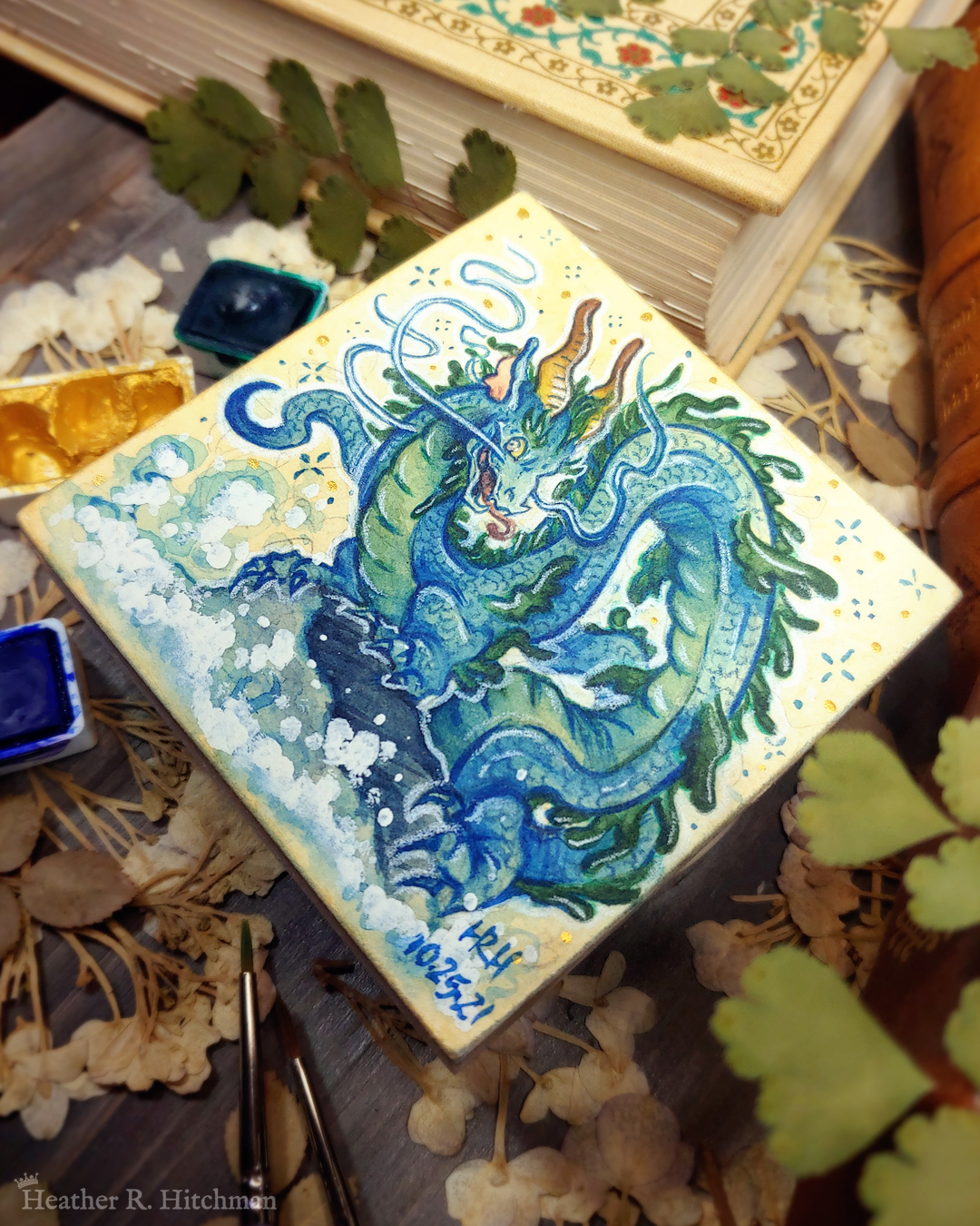 Photograph of a watercolor and gouache painting of an evil rotten leviathan blue water dragon. The dragon has a long, snake like blue and green body with no wings. His body coils across the 3x3 paper against a soft sepia background filled with small gold and blue stars. He is standing on a rock with waves crashing up against it. The dragon’s face has 2 long whiskers that float up from his cheeks to above his head. He has golden goat like horns that twist up into the air. The dragon’s torso is facing the viewer, his face is in profile as he looks towards the viewer with a menacing grin. The painting sits on top of a table covered with watercolor paint pans, books, and foliage in the background.