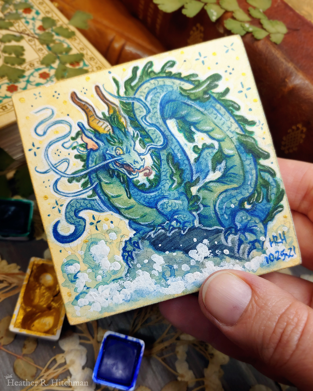 Photograph of a watercolor and gouache painting of an evil rotten leviathan blue water dragon. The dragon has a long, snake like blue and green body with no wings. His body coils across the 3x3 paper against a soft sepia background filled with small gold and blue stars. He is standing on a rock with waves crashing up against it. The dragon’s face has 2 long whiskers that float up from his cheeks to above his head. He has golden goat like horns that twist up into the air. The dragon’s torso is facing the viewer, his face is in profile as he looks towards the viewer with a menacing grin. The artist’s hand is holding the small painting over top of a table covered with watercolor paint pans, books, and foliage in the background.