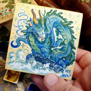 Photograph of a watercolor and gouache painting of an evil rotten leviathan blue water dragon. The dragon has a long, snake like blue and green body with no wings. His body coils across the 3x3 paper against a soft sepia background filled with small gold and blue stars. He is standing on a rock with waves crashing up against it. The dragon’s face has 2 long whiskers that float up from his cheeks to above his head. He has golden goat like horns that twist up into the air. The dragon’s torso is facing the viewer, his face is in profile as he looks towards the viewer with a menacing grin. The artist’s hand is holding the small painting over top of a table covered with watercolor paint pans, books, and foliage in the background.