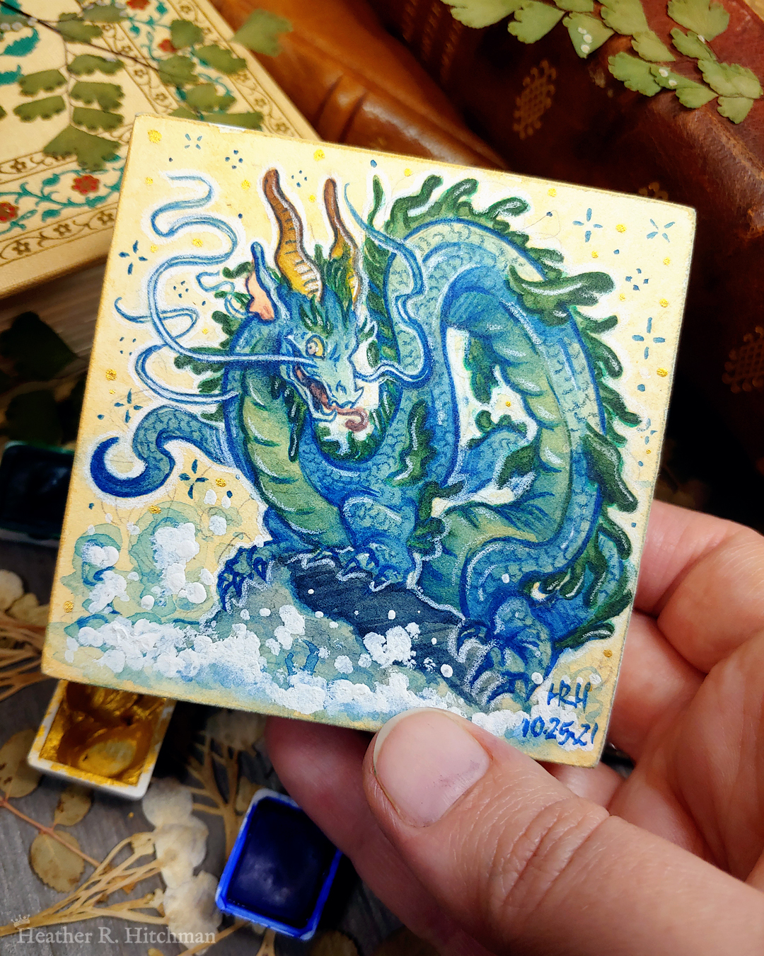 Photograph of a watercolor and gouache painting of an evil rotten leviathan blue water dragon. The dragon has a long, snake like blue and green body with no wings. His body coils across the 3x3 paper against a soft sepia background filled with small gold and blue stars. He is standing on a rock with waves crashing up against it. The dragon’s face has 2 long whiskers that float up from his cheeks to above his head. He has golden goat like horns that twist up into the air. The dragon’s torso is facing the viewer, his face is in profile as he looks towards the viewer with a menacing grin. The artist’s hand is holding the small painting over top of a table covered with watercolor paint pans, books, and foliage in the background.