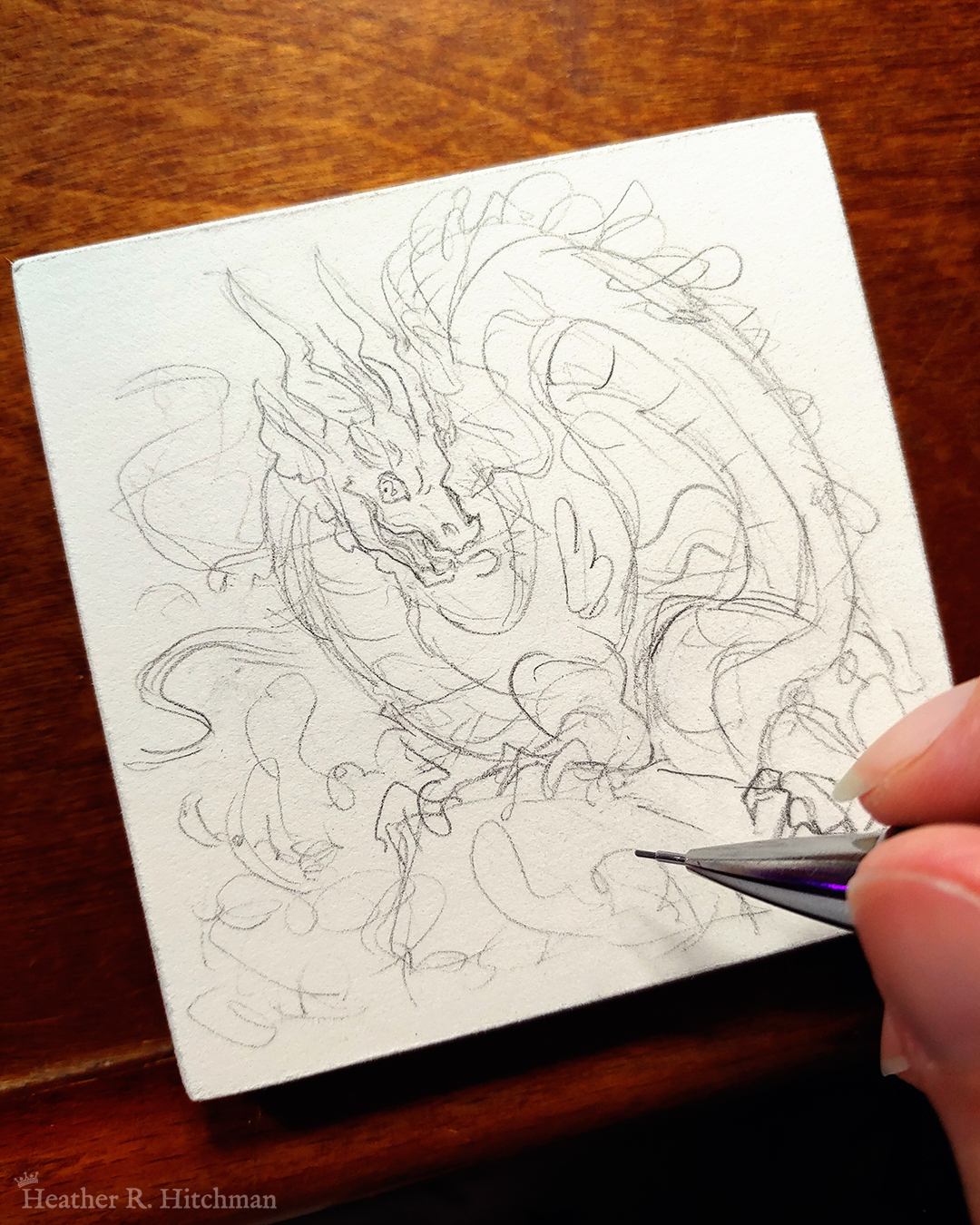 Photograph of a graphite sketch of an evil rotten leviathan water dragon. The dragon has a long, snake like body with no wings. His body coils across the 3x3 paper. He is standing on a rock with waves crashing up against it. The dragon’s face has 2 long whiskers that float up from his cheeks to above his head. He has goat like horns that twist up into the air. The dragon’s torso is facing the viewer, his face is in profile as he looks towards the viewer with a menacing grin. The artist’s hand holds a mechanical pencil over the drawing on the bottom right of the photograph.