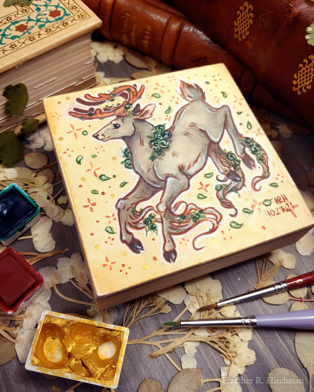 Photograph of a watercolor and gouache painting of a jumping gray brown stag. His body is tilting downwards to the left with his head upright. The background is a soft sepia wash of color filled with small brown stars and green leaves circling the stag. The stag has small green roses around his neck, in his antlers, and falling from his hooves. His face is in profile as he happily looks off into the distance. The artist’s hand is holding the small painting over top of a table covered with watercolor paint pans, books, and foliage in the background. The painting sits on top of a table covered with watercolor paint pans, books, and foliage in the background.