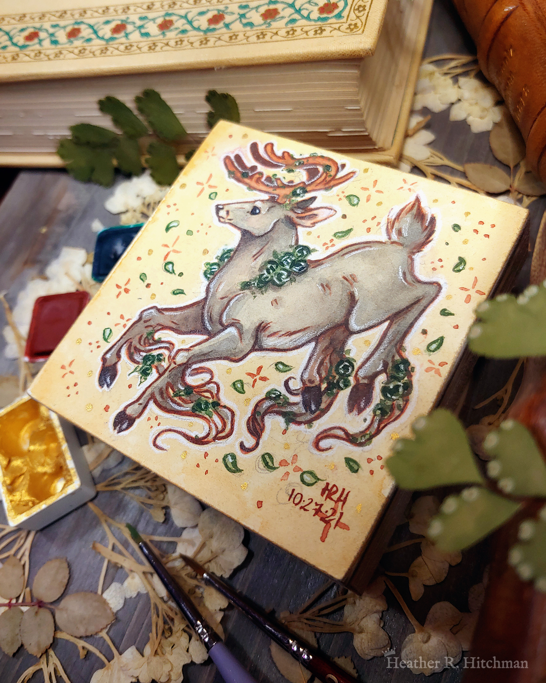 Photograph of a watercolor and gouache painting of a jumping gray brown stag. His body is tilting downwards to the left with his head upright. The background is a soft sepia wash of color filled with small brown stars and green leaves circling the stag. The stag has small green roses around his neck, in his antlers, and falling from his hooves. His face is in profile as he happily looks off into the distance. The artist’s hand is holding the small painting over top of a table covered with watercolor paint pans, books, and foliage in the background. The painting sits on top of a table covered with watercolor paint pans, books, and foliage in the background.
