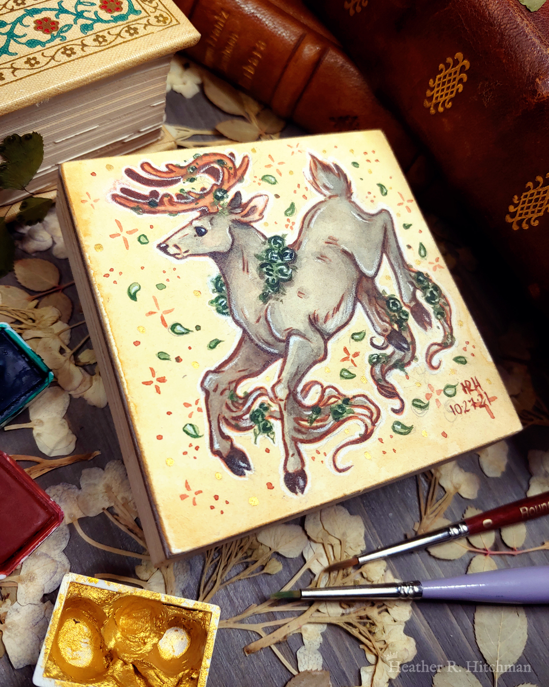 Photograph of a watercolor and gouache painting of a jumping gray brown stag. His body is tilting downwards to the left with his head upright. The background is a soft sepia wash of color filled with small brown stars and green leaves circling the stag. The stag has small green roses around his neck, in his antlers, and falling from his hooves. His face is in profile as he happily looks off into the distance. The artist’s hand is holding the small painting over top of a table covered with watercolor paint pans, books, and foliage in the background. The painting sits on top of a table covered with watercolor paint pans, books, and foliage in the background.