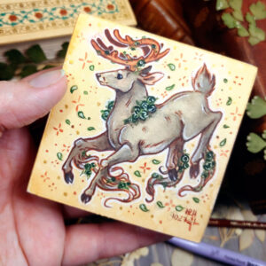 Photograph of a watercolor and gouache painting of a jumping gray brown stag. His body is tilting downwards to the left with his head upright. The background is a soft sepia wash of color filled with small brown stars and green leaves circling the stag. The stag has small green roses around his neck, in his antlers, and falling from his hooves. His face is in profile as he happily looks off into the distance. The artist’s hand is holding the small painting over top of a table covered with watercolor paint pans, books, and foliage in the background.