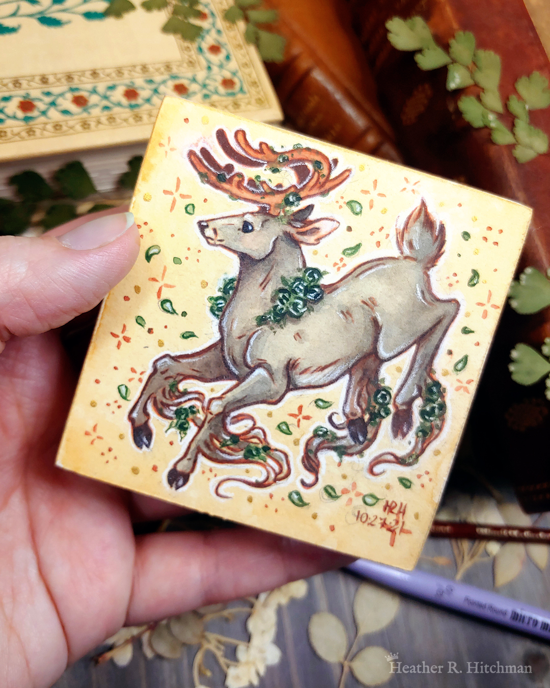 Photograph of a watercolor and gouache painting of a jumping gray brown stag. His body is tilting downwards to the left with his head upright. The background is a soft sepia wash of color filled with small brown stars and green leaves circling the stag. The stag has small green roses around his neck, in his antlers, and falling from his hooves. His face is in profile as he happily looks off into the distance. The artist’s hand is holding the small painting over top of a table covered with watercolor paint pans, books, and foliage in the background.