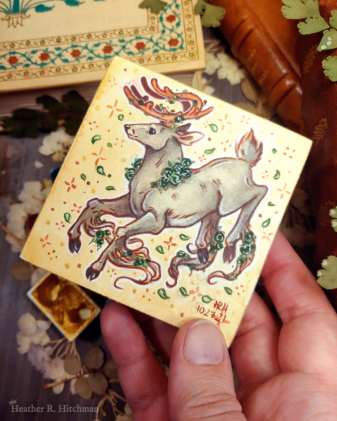Photograph of a watercolor and gouache painting of a jumping gray brown stag. His body is tilting downwards to the left with his head upright. The background is a soft sepia wash of color filled with small brown stars and green leaves circling the stag. The stag has small green roses around his neck, in his antlers, and falling from his hooves. His face is in profile as he happily looks off into the distance. The artist’s hand is holding the small painting over top of a table covered with watercolor paint pans, books, and foliage in the background.