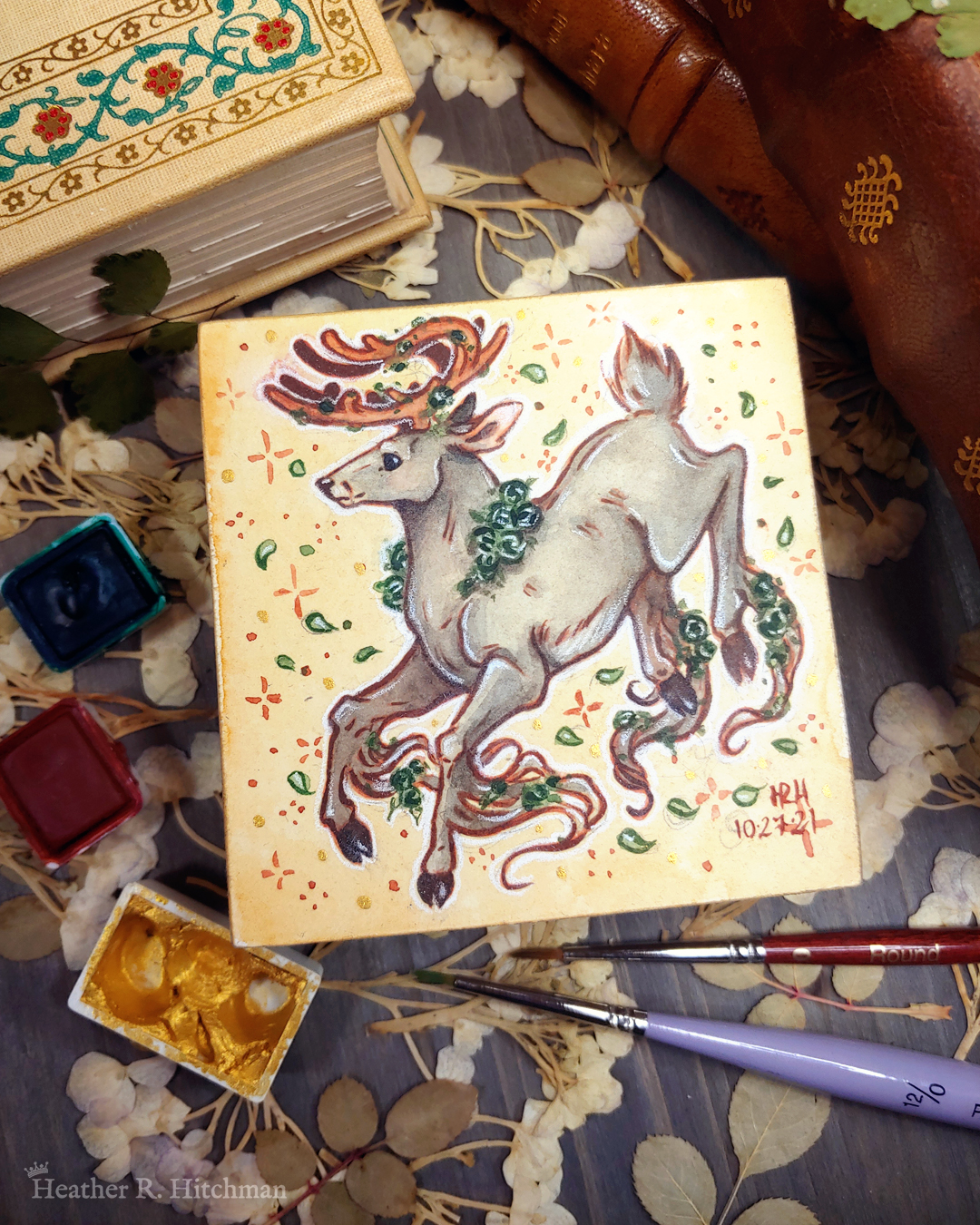 Photograph of a watercolor and gouache painting of a jumping gray brown stag. His body is tilting downwards to the left with his head upright. The background is a soft sepia wash of color filled with small brown stars and green leaves circling the stag. The stag has small green roses around his neck, in his antlers, and falling from his hooves. His face is in profile as he happily looks off into the distance. The artist’s hand is holding the small painting over top of a table covered with watercolor paint pans, books, and foliage in the background. The painting sits on top of a table covered with watercolor paint pans, books, and foliage in the background.