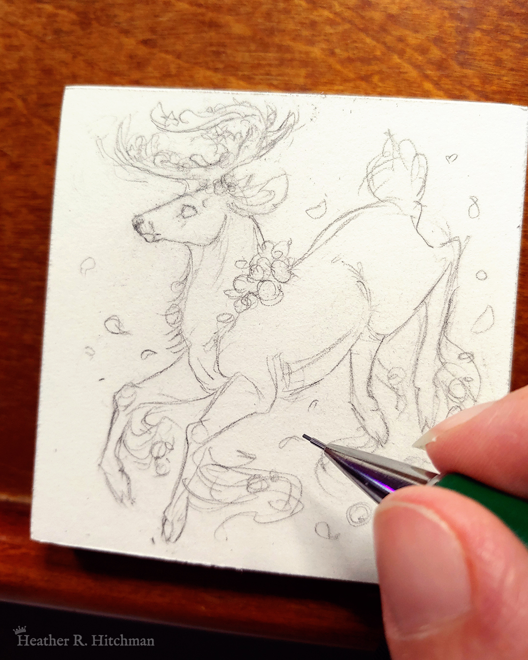 Photograph of a graphite sketch of a jumping gray brown stag. His body is tilting downwards to the left with his head upright. The background is white with a few small leaves circling the stag. The stag has small roses around his neck, in his antlers, and falling from his hooves. His face is in profile as he happily looks off into the distance. The artist’s hand holds a mechanical pencil over the drawing on the bottom right of the photograph.