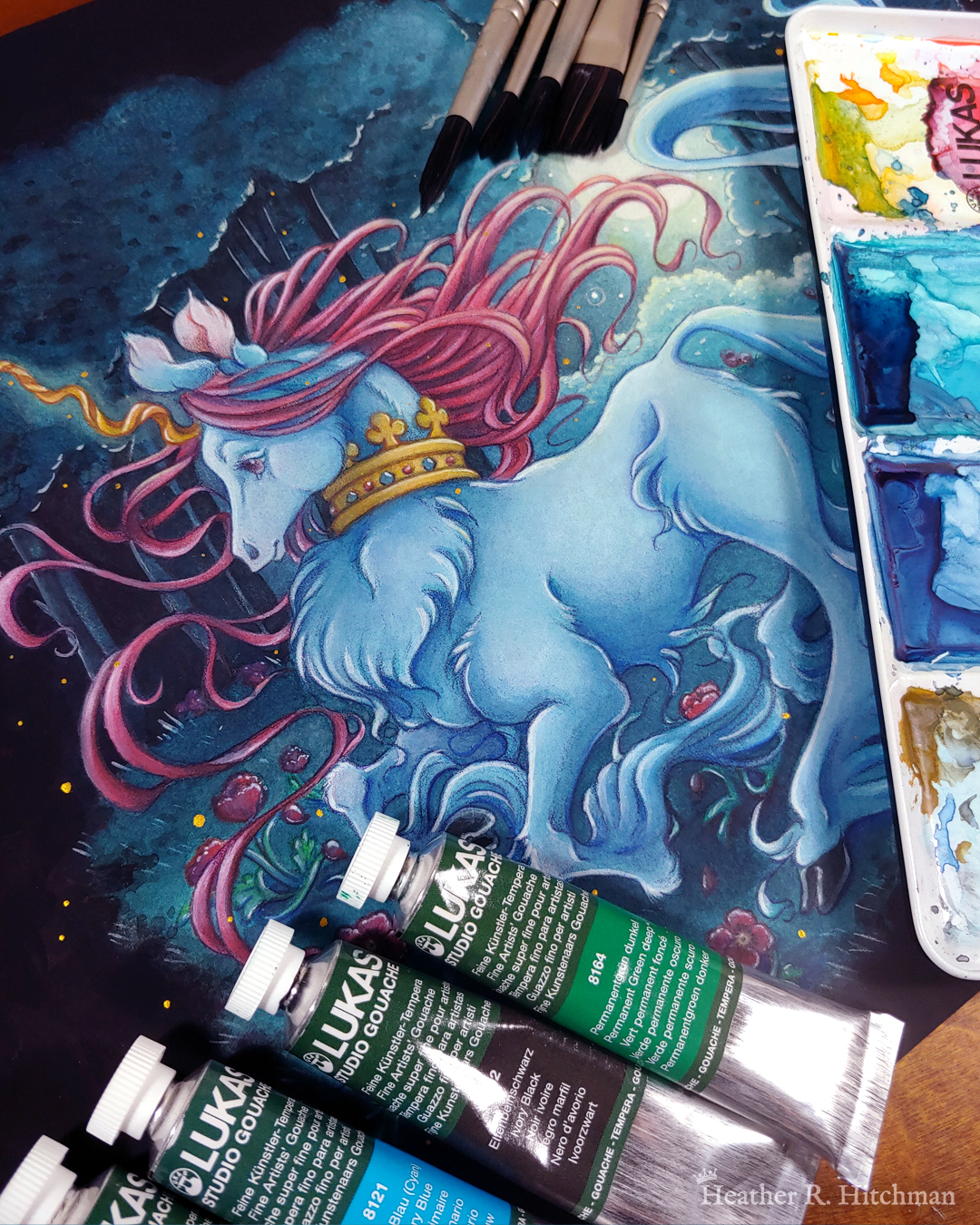 Photograph of a full body watercolor, gouache, and colored pencil painting of a unicorn running through a moonlit forest. The unicorn has blue fur that flows in elegant tufts from their chest, ankles, and lion-like tail. He has a mane of raspberry red that swirls around in the wind behind the unicorn’s head. The unicorn has a twisting golden horn, and wears a golden crown encrusted with red and blue jewels around their neck. Running in profile to the left with their front hooves close to their chest, the unicorn has a peaceful expression on its face. The background is a cool greenish blue night forest scene, that is surrounded by black. In the field red poppies bloom under the unicorn’s hooves. Trees with dark branches are in the far background, with an opening behind the unicorn that reveals a large full moon glowing white. The scene is sprinkled with tiny golden orbs of light. Tubes of watercolor paint rest at the bottom left of the painting in a diagonal. A watercolor pallet is partially visible over the far right side of the painting. Watercolor brushes are lying on the painting at the top of the photograph, only the tips are visible.