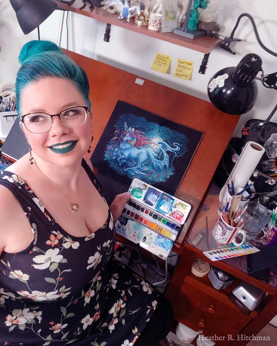 Photograph of the artist Heather R. Hitchman in her art studio sitting at her art desk. She is holding a watercolor pallet in her left hand and looking off camera to the left with a smile. On her art desk is a full body watercolor, gouache, and colored pencil painting of a unicorn running through a moonlit forest. The unicorn has blue fur that flows in elegant tufts from their chest, ankles, and lion-like tail. He has a mane of raspberry red that swirls around in the wind behind the unicorn’s head. The unicorn has a twisting golden horn, and wears a golden crown encrusted with red and blue jewels around their neck. Running in profile to the left with their front hooves close to their chest, the unicorn has a peaceful expression on its face. The background is a cool greenish blue night forest scene, that is surrounded by black. In the field red poppies bloom under the unicorn’s hooves. Trees with dark branches are in the far background, with an opening behind the unicorn that reveals a large full moon glowing white. The scene is sprinkled with tiny golden orbs of light.