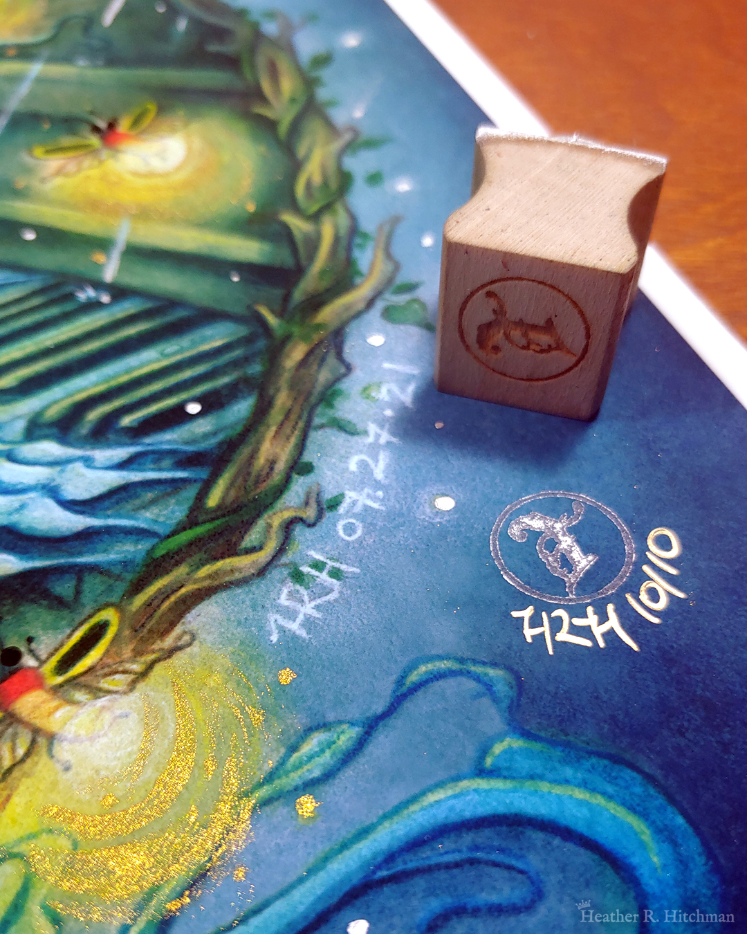 Close up photograph of a watercolor and gouache painting print of “Blue Hair and the Piano” by Heather R. Hitchman. The photograph focuses on the bottom right corner of the painting, where part of a branch wreath and glowing fireflies are visible. The back sides of the fire flies has been embellished with golden paint and shimmers in the light. There is a silver stamp of the Terratoff icon in the corner, which has been signed and numbered in gold ink. On top of the print the stamp is resting on it’s side.