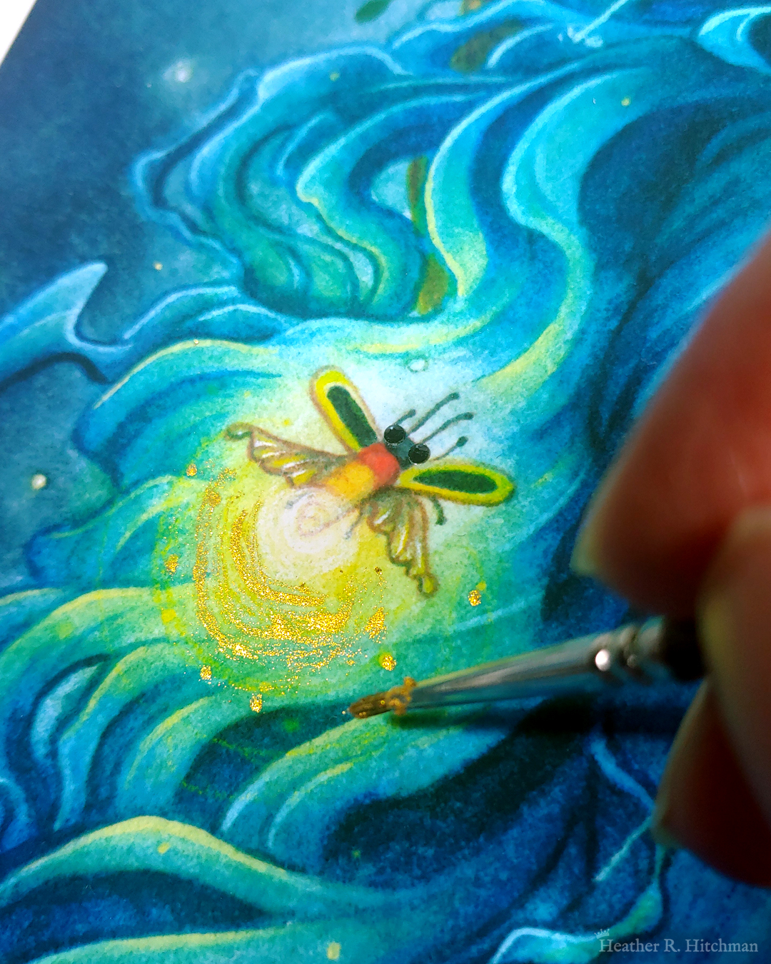 Close up photograph of a watercolor and gouache painting print of “Blue Hair and the Piano” by Heather R. Hitchman. The painting has a moody and rainy atmosphere. The photograph focuses on the firefly hovering over the faun’s flowing blue hair. The firefly has shimmering black crystals for eyes, and a bright glowing light on it’s bottom. The fingertips of the artist are on the right side of the photograph holding a tiny paintbrush as she paints golden metallic paint on the firefly’s light, creating a shimmering orb effect.