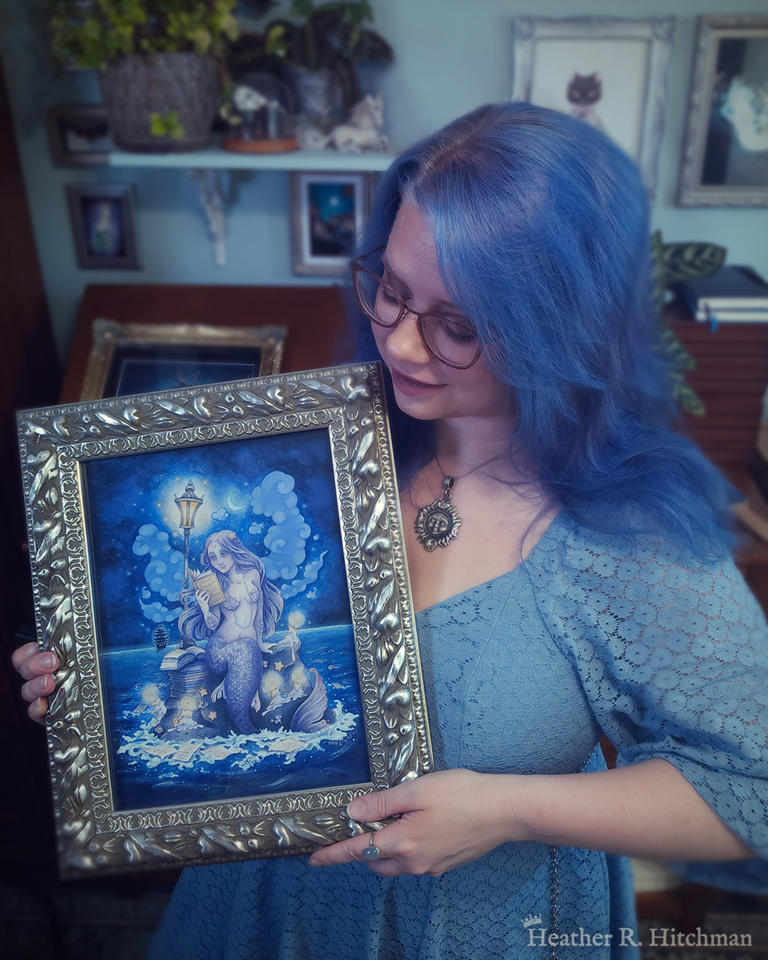 Photograph of the artist Heather R. Hitchman holding a framed watercolor and gouache full body painting of a purple mermaid reading a book under the moonlight while sitting on a gray rock in the middle of the sea. The rock is covered in glowing candles and also has a pile of books besides the mermaid’s tail. An old fashioned street lantern covered in barnacles is attached to the rock, illuminating orange light against a dark blue night sky. Gentle blue waves wash up against the rock. Loose white pages from books float in the water into the distance, leading towards a mysterious black ship that vanishes into the night. Puffy light blue fish shaped clouds fill the nigh sky surrounding a crescent moon. The mermaid is holding a book in her right hand up to her face. Her other hand is placed behind her as she leans back slightly, her left arm supporting her weight. Her body faces the viewer, and she has a peaceful smile as she looks towards the book and reads. Heather is standing in front of a wooden art desk in her studio filled with potted plants and artwork.