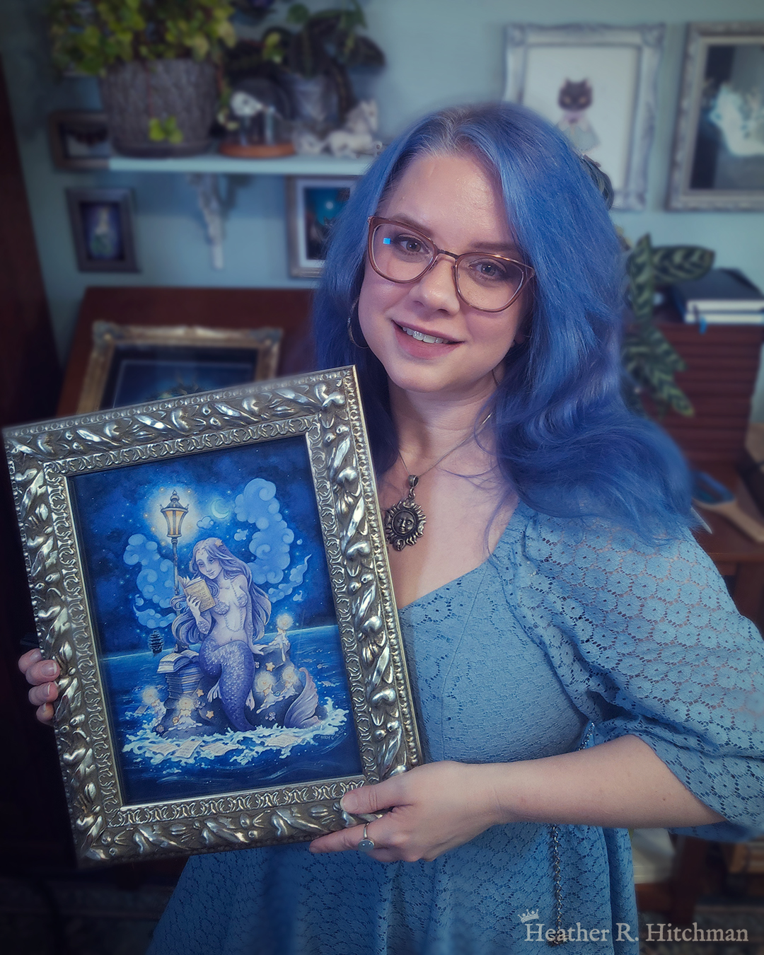 Photograph of the artist Heather R. Hitchman holding a framed watercolor and gouache full body painting of a purple mermaid reading a book under the moonlight while sitting on a gray rock in the middle of the sea. The rock is covered in glowing candles and also has a pile of books besides the mermaid’s tail. An old fashioned street lantern covered in barnacles is attached to the rock, illuminating orange light against a dark blue night sky. Gentle blue waves wash up against the rock. Loose white pages from books float in the water into the distance, leading towards a mysterious black ship that vanishes into the night. Puffy light blue fish shaped clouds fill the nigh sky surrounding a crescent moon. The mermaid is holding a book in her right hand up to her face. Her other hand is placed behind her as she leans back slightly, her left arm supporting her weight. Her body faces the viewer, and she has a peaceful smile as she looks towards the book and reads. Heather is standing in front of a wooden art desk in her studio filled with potted plants and artwork.