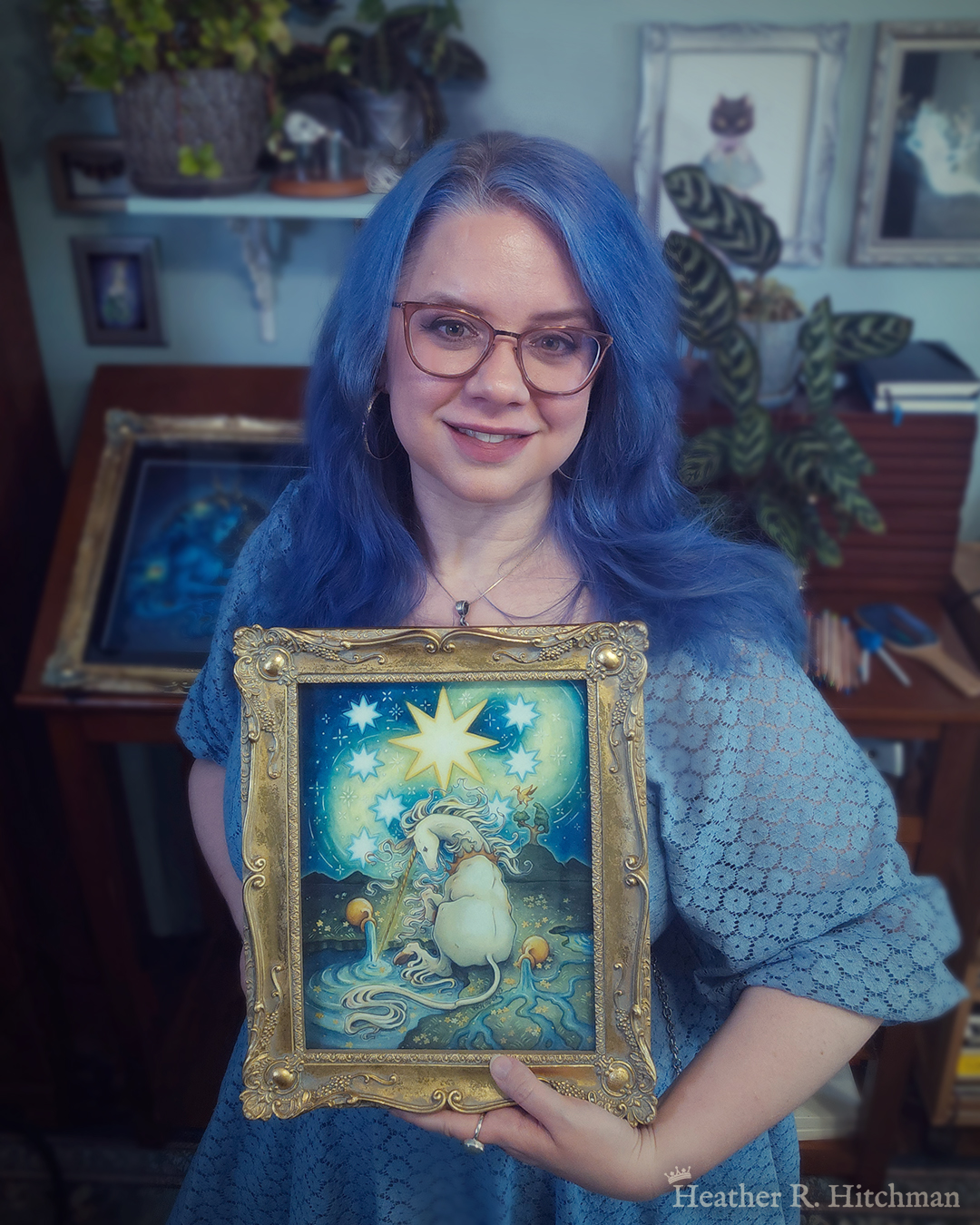 Photograph of the artist Heather R. Hitchman holding a framed watercolor and gouache painting of a unicorn sitting by a lake under a night sky. The sky is dark blue and filled with tiny white stars. Seven large white stars surround a massive yellow star in the center of the sky. A swirl of bright yellow light weaves upwards towards the heavens, illuminating the scene below. The unicorn sits in front of a landscape scene, with green mountaintops in the background. On one of the mountaintops a small tree grows, on top of the branches a yellow ibis bird flaps its wings. The unicorn is sitting with its back towards the viewer, the head turned in profile as she dips her long golden horn into the lake to the left of the image. The unicorn wears a golden crown around her neck. Two golden urns rest on their sides on the ground, spilling water onto the grassy scene that trickles into the lake waters. Heather is standing in front of a wooden art desk in her studio filled with potted plants and artwork.