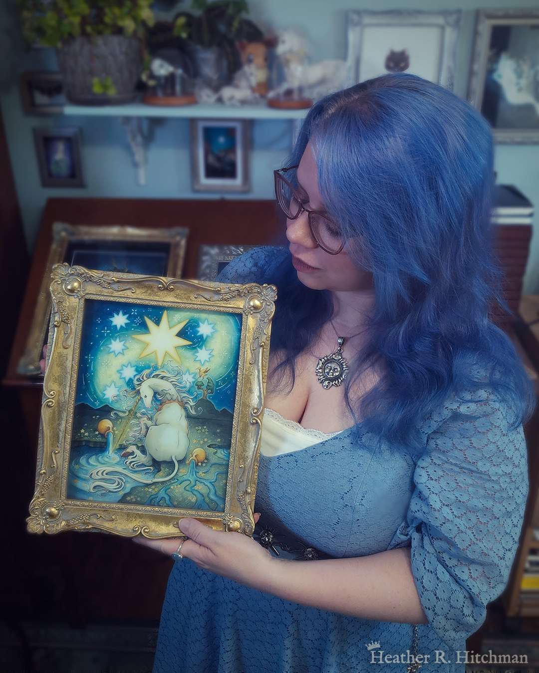 Photograph of the artist Heather R. Hitchman holding a framed watercolor and gouache painting of a unicorn sitting by a lake under a night sky. The sky is dark blue and filled with tiny white stars. Seven large white stars surround a massive yellow star in the center of the sky. A swirl of bright yellow light weaves upwards towards the heavens, illuminating the scene below. The unicorn sits in front of a landscape scene, with green mountaintops in the background. On one of the mountaintops a small tree grows, on top of the branches a yellow ibis bird flaps its wings. The unicorn is sitting with its back towards the viewer, the head turned in profile as she dips her long golden horn into the lake to the left of the image. The unicorn wears a golden crown around her neck. Two golden urns rest on their sides on the ground, spilling water onto the grassy scene that trickles into the lake waters. Heather is standing in front of a wooden art desk in her studio filled with potted plants and artwork.