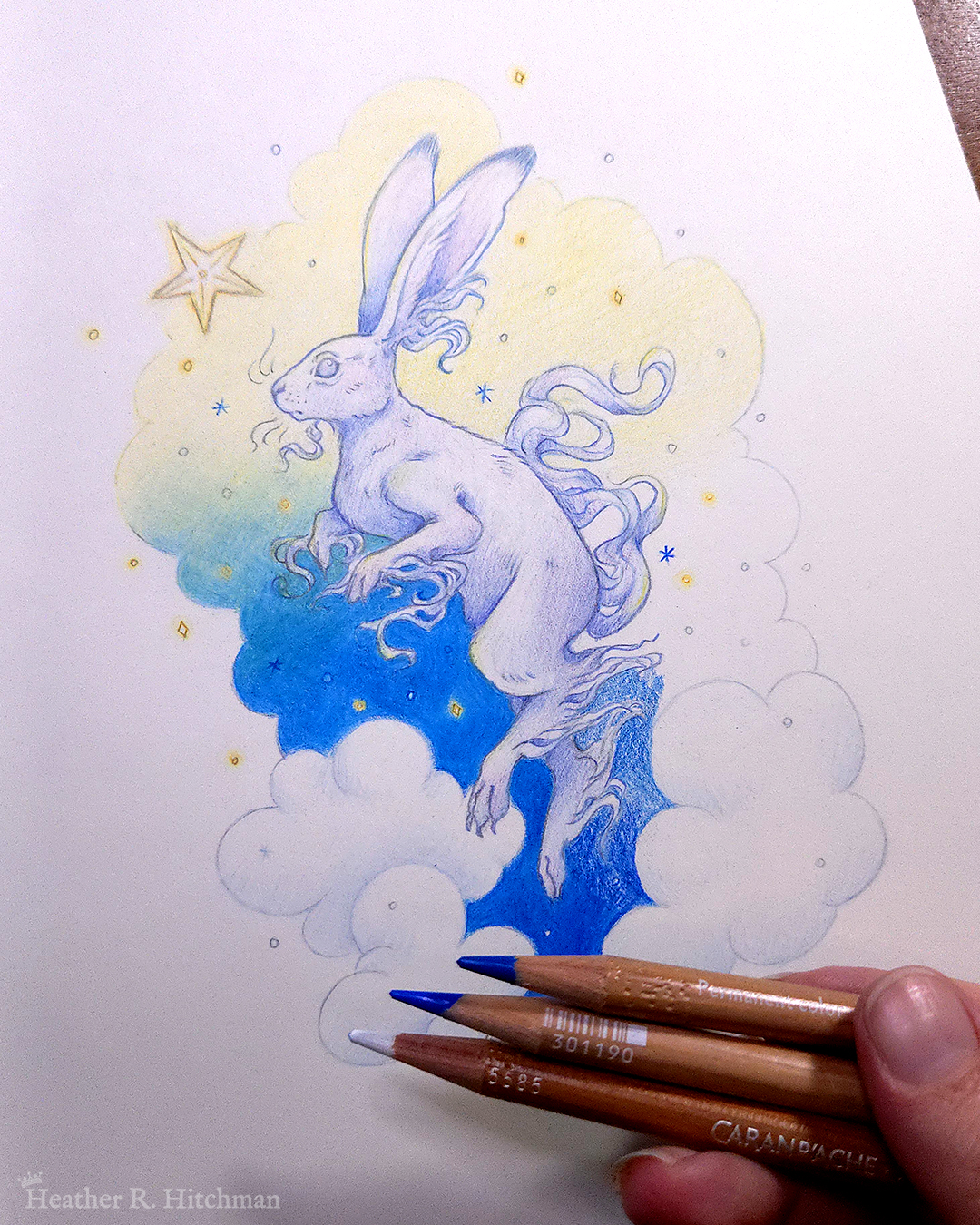 Process photograph of a colored pencil and graphite drawing of a white yule hare with wispy long fur coming from it’s ears, paws, and tail. The hare is floating in profile facing to the left, hovering over 3 large clouds against the gradient of a blue and yellow night sky in the background. The gradient of the sky is in a cloudy shape that frames the clouds and the yule hare. Tiny yellow stars and white snowflakes are scattered around the scene. The hare's head is looking up towards a bright yellow star to the left with a stoic expression. The hand of the artist is to the bottom right holding 3 pencils over the image.