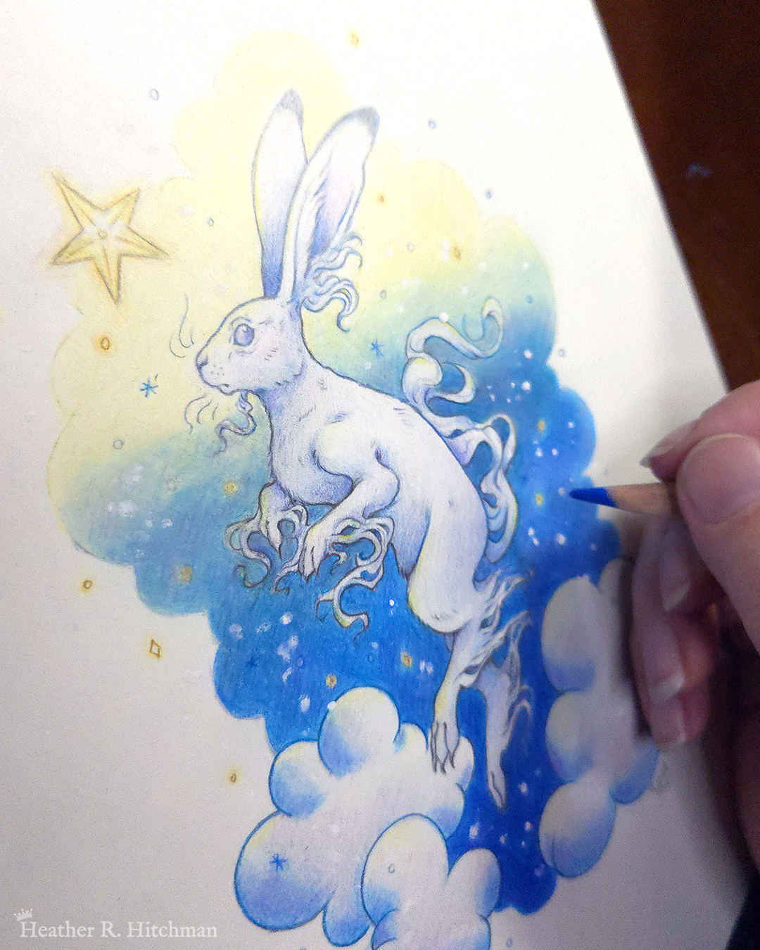 Process photograph of a colored pencil and graphite drawing of a white yule hare with wispy long fur coming from it’s ears, paws, and tail. The hare is floating in profile facing to the left, hovering over 3 large clouds against the gradient of a blue and yellow night sky in the background. The gradient of the sky is in a cloudy shape that frames the clouds and the yule hare. Tiny yellow stars and white snowflakes are scattered around the scene. The hare's head is looking up towards a bright yellow star to the left with a stoic expression. The hand of the artist is to the right holding a pencil over the image.
