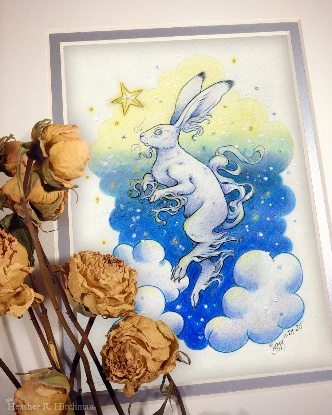 Photograph of a matted print of a colored pencil and graphite drawing of a white yule hare with wispy long fur coming from it’s ears, paws, and tail. The hare is floating in profile facing to the left, hovering over 3 large clouds against the gradient of a blue and yellow night sky in the background. The gradient of the sky is in a cloudy shape that frames the clouds and the yule hare. Tiny yellow stars and white snowflakes are scattered around the scene. The hare's head is looking up towards a bright yellow star to the left with a stoic expression. The mat-board is white on the top layer and silver on the second layer. Yellow roses rest on top of the print to the left. A small silver stamp with the Terratoff icon is at the bottom left corner of the print, under the roses.