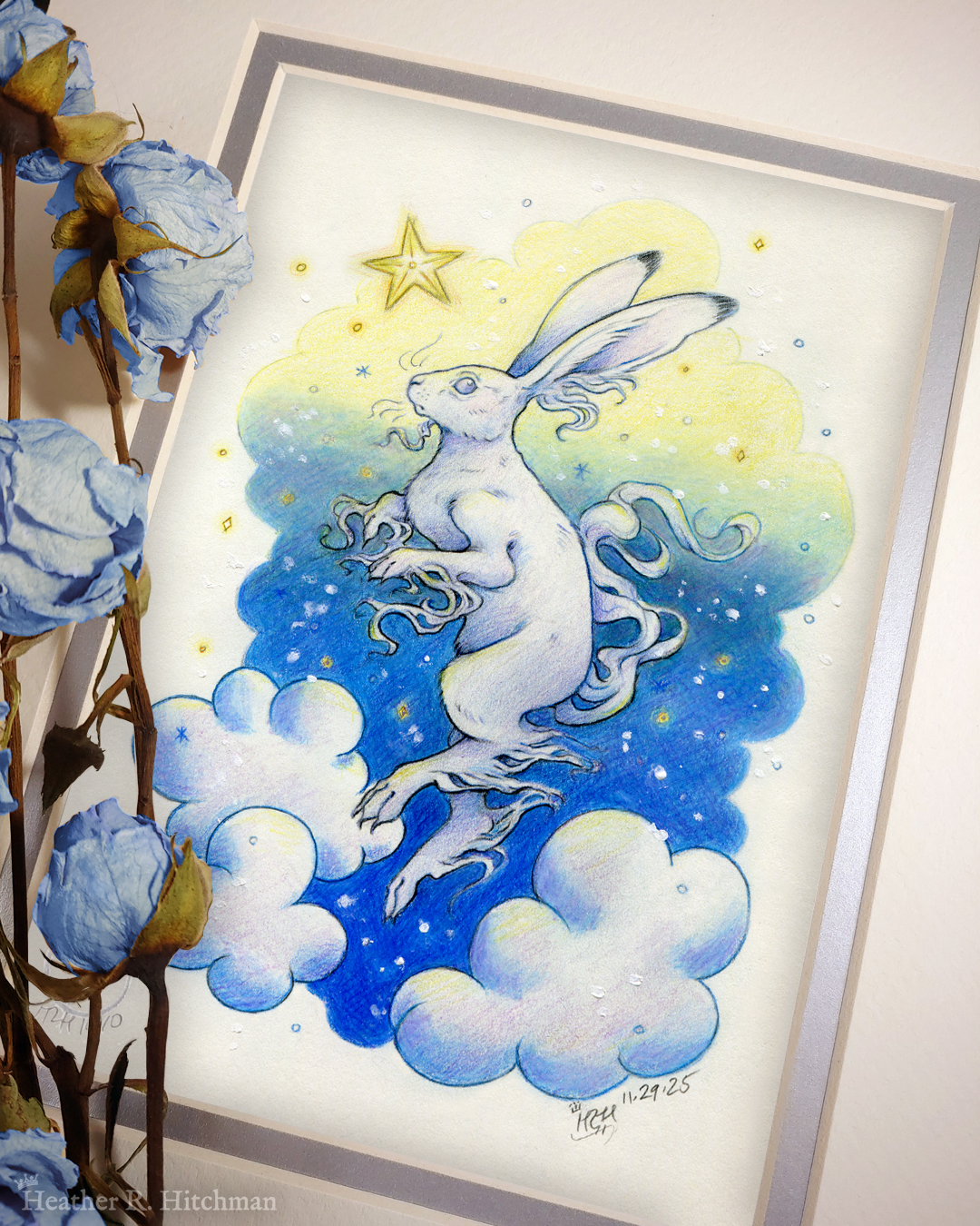 Photograph of a matted print of a colored pencil and graphite drawing of a white yule hare with wispy long fur coming from it’s ears, paws, and tail. The hare is floating in profile facing to the left, hovering over 3 large clouds against the gradient of a blue and yellow night sky in the background. The gradient of the sky is in a cloudy shape that frames the clouds and the yule hare. Tiny yellow stars and white snowflakes are scattered around the scene. The hare's head is looking up towards a bright yellow star to the left with a stoic expression. The mat-board is white on the top layer and silver on the second layer. Blue roses rest on top of the print to the left. A small silver stamp with the Terratoff icon is at the bottom left corner of the print, under the roses.