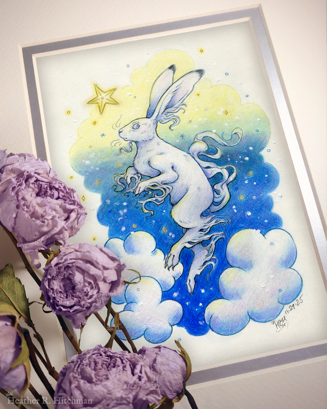 Photograph of a matted print of a colored pencil and graphite drawing of a white yule hare with wispy long fur coming from it’s ears, paws, and tail. The hare is floating in profile facing to the left, hovering over 3 large clouds against the gradient of a blue and yellow night sky in the background. The gradient of the sky is in a cloudy shape that frames the clouds and the yule hare. Tiny yellow stars and white snowflakes are scattered around the scene. The hare's head is looking up towards a bright yellow star to the left with a stoic expression. The mat-board is white on the top layer and silver on the second layer. Purple roses rest on top of the print to the left. A small silver stamp with the Terratoff icon is at the bottom left corner of the print, under the roses.