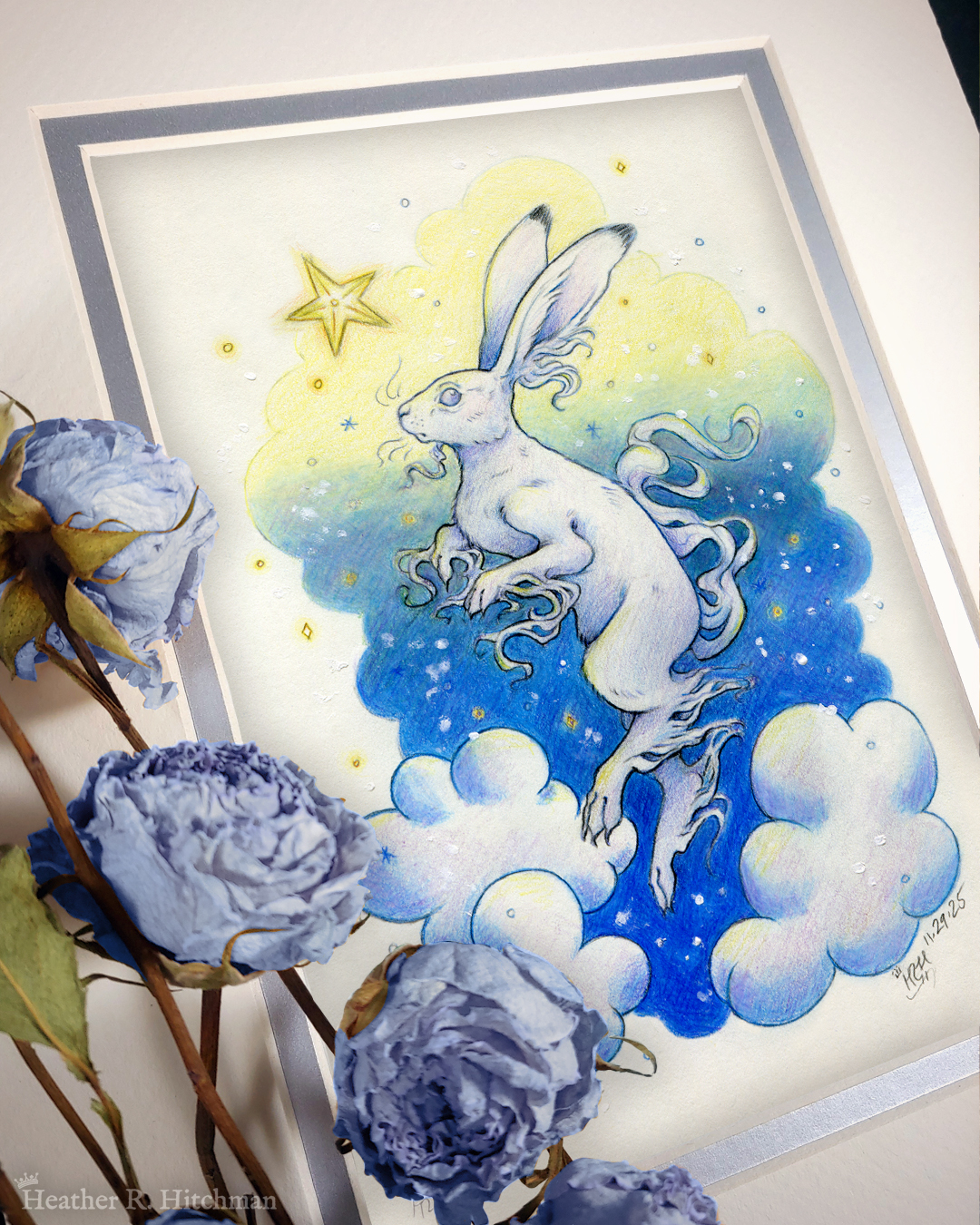 Photograph of a matted print of a colored pencil and graphite drawing of a white yule hare with wispy long fur coming from it’s ears, paws, and tail. The hare is floating in profile facing to the left, hovering over 3 large clouds against the gradient of a blue and yellow night sky in the background. The gradient of the sky is in a cloudy shape that frames the clouds and the yule hare. Tiny yellow stars and white snowflakes are scattered around the scene. The hare's head is looking up towards a bright yellow star to the left with a stoic expression. The mat-board is white on the top layer and silver on the second layer. Blue roses rest on top of the print to the left. A small silver stamp with the Terratoff icon is at the bottom left corner of the print, under the roses.
