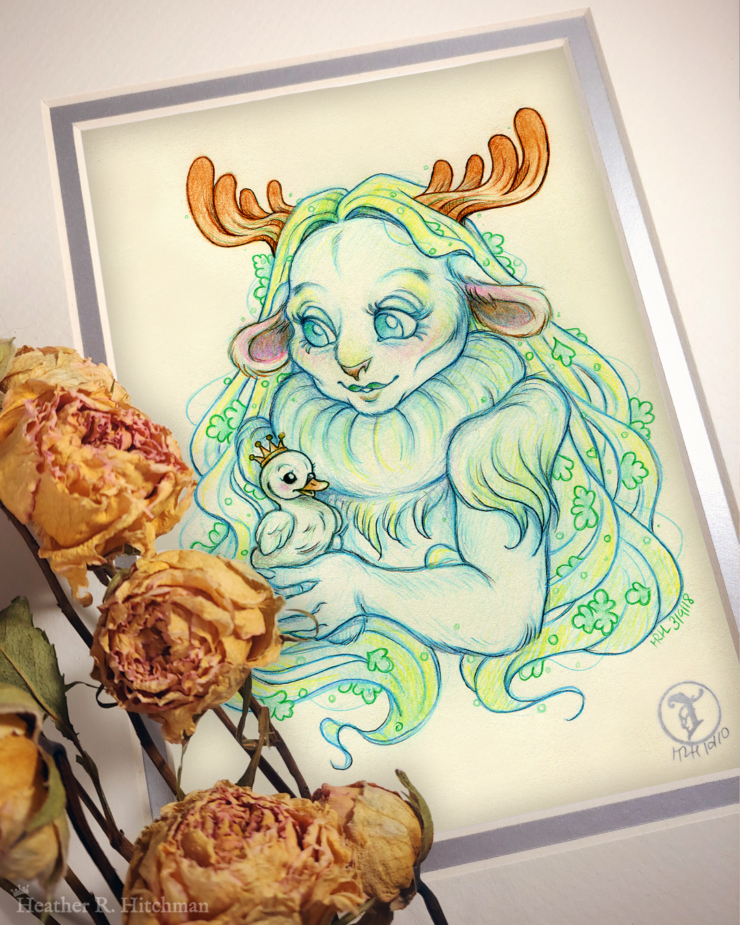 Photograph of a matted print of a graphite and colored pencil chest up portrait drawing of a child female faun with soft blueish green fur, large blue eyes, and small moose antlers. The faun’s fur is gathered into a poofy collar around her neck, creating the illusion of a ruff. The fur gathered at her shoulders looks similar to shoulder pads. She is posing at a ¾ angle to the left holding a tiny swan gosling in her hands. The gosling is wearing a tiny gold crown on its head and is looking up at the faun adoringly. The faun looks back down at the gosling, blushing with a smile on her face. Her long straight blond hair cascades around her back and shoulders, decorated with small green fern leaves. The mat-board is white on the top layer and silver on the second layer. Yellow roses rest on top of the print to the left. A small silver stamp with the Terratoff icon is at the bottom right corner of the print.