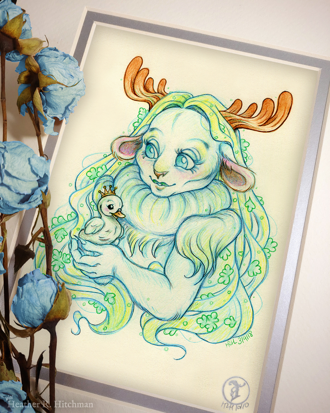 Photograph of a matted print of a graphite and colored pencil chest up portrait drawing of a child female faun with soft blueish green fur, large blue eyes, and small moose antlers. The faun’s fur is gathered into a poofy collar around her neck, creating the illusion of a ruff. The fur gathered at her shoulders looks similar to shoulder pads. She is posing at a ¾ angle to the left holding a tiny swan gosling in her hands. The gosling is wearing a tiny gold crown on its head and is looking up at the faun adoringly. The faun looks back down at the gosling, blushing with a smile on her face. Her long straight blond hair cascades around her back and shoulders, decorated with small green fern leaves. The mat-board is white on the top layer and silver on the second layer. Blue roses rest on top of the print to the left. A small silver stamp with the Terratoff icon is at the bottom right corner of the print.