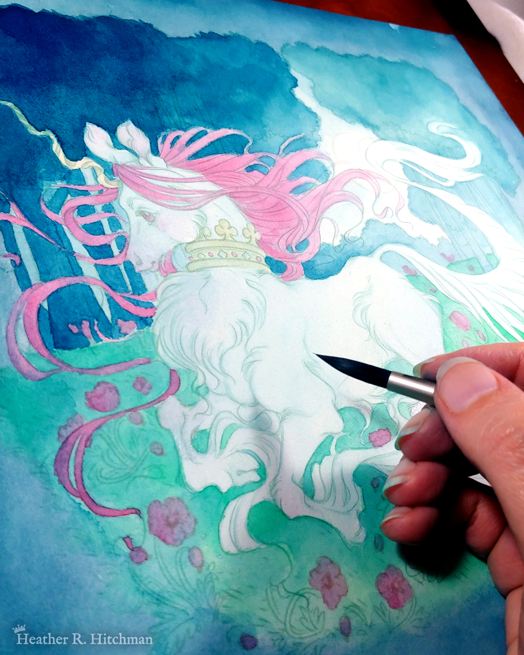 Process photograph of a full body watercolor, gouache, and colored pencil painting of a unicorn running through a moonlit forest. The artist’s hand is resting on the bottom right section of the painting holding a paintbrush. The painting is very loose and sketchy with soft layers of watercolors and colored pencils created so far. The unicorn has light blue fur that flows in elegant tufts from their chest, ankles, and lion-like tail. He has a mane of raspberry red that swirls around in the wind behind the unicorn’s head. The unicorn has a twisting golden horn, and wears a golden crown encrusted with red and blue jewels around their neck. Running in profile to the left with their front hooves close to their chest, the unicorn has a peaceful expression on its face. The background is a cool greenish blue night forest scene. In the field red poppies bloom under the unicorn’s hooves. Trees with dark branches are in the far background, with an opening behind the unicorn that reveals a large full moon glowing white.