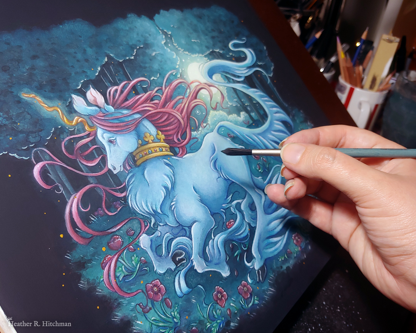 Photograph of a full body watercolor, gouache, and colored pencil painting of a unicorn running through a moonlit forest. The unicorn has blue fur that flows in elegant tufts from their chest, ankles, and lion-like tail. He has a mane of raspberry red that swirls around in the wind behind the unicorn’s head. The unicorn has a twisting golden horn, and wears a golden crown encrusted with red and blue jewels around their neck. Running in profile to the left with their front hooves close to their chest, the unicorn has a peaceful expression on its face. The background is a cool greenish blue night forest scene, that is surrounded by black. In the field red poppies bloom under the unicorn’s hooves. Trees with dark branches are in the far background, with an opening behind the unicorn that reveals a large full moon glowing white. The scene is sprinkled with tiny golden orbs of light. The painting is at an angle on top of an art desk, the artist’s hand is resting on top of the painting to the right holding a paintbrush.