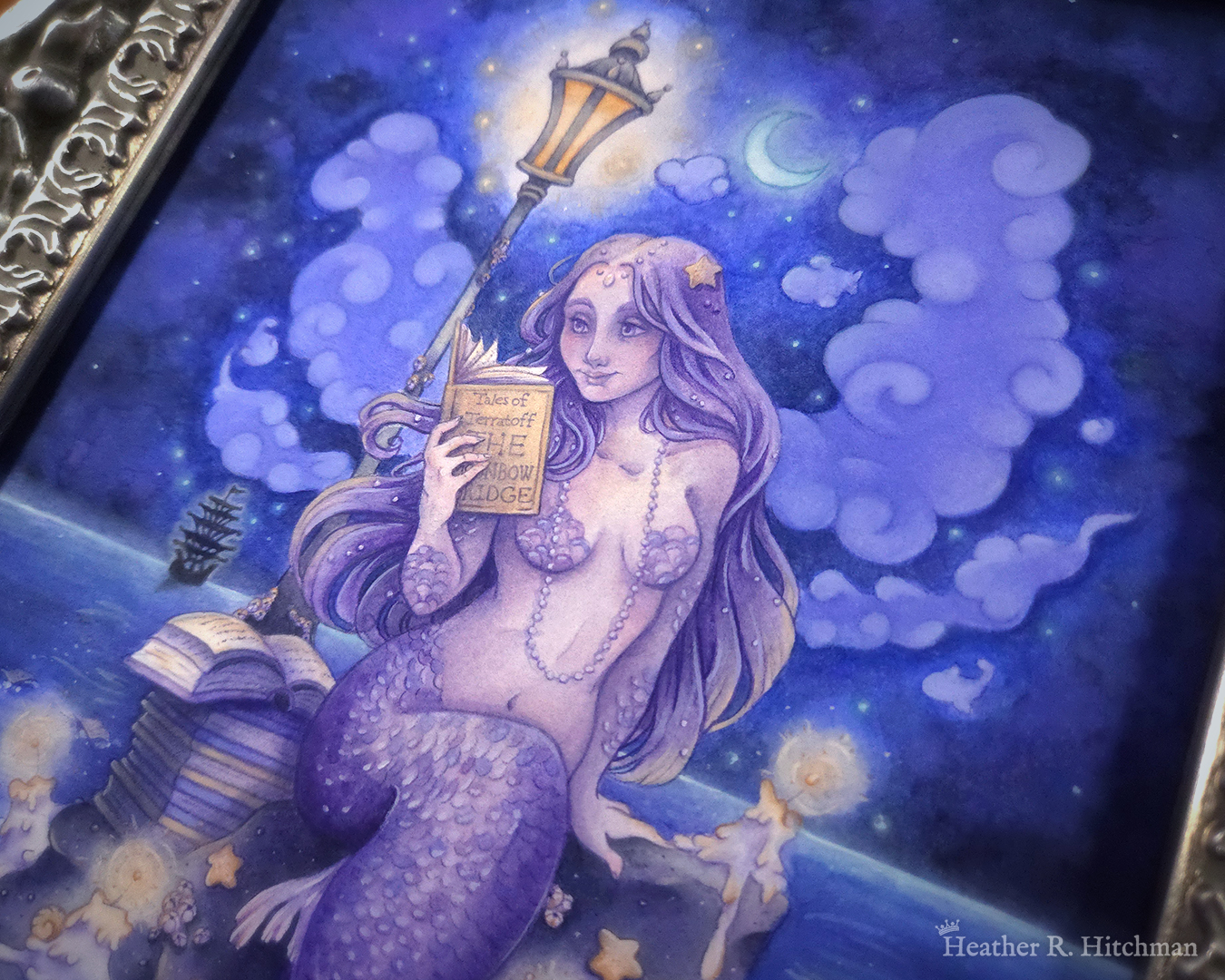 Close up of a watercolor and gouache full body painting of a purple mermaid reading a book under the moonlight while sitting on a gray rock in the middle of the sea. An old fashioned street lantern covered in barnacles is attached to the rock, illuminating orange light against a dark blue night sky. The mermaid is holding a book in her right hand up to her face. Her other hand is placed behind her as she leans back slightly, her left arm supporting her weight. Her body faces the viewer, and she has a peaceful smile as she looks towards the book and reads. The painting is in a silver ornate frame.