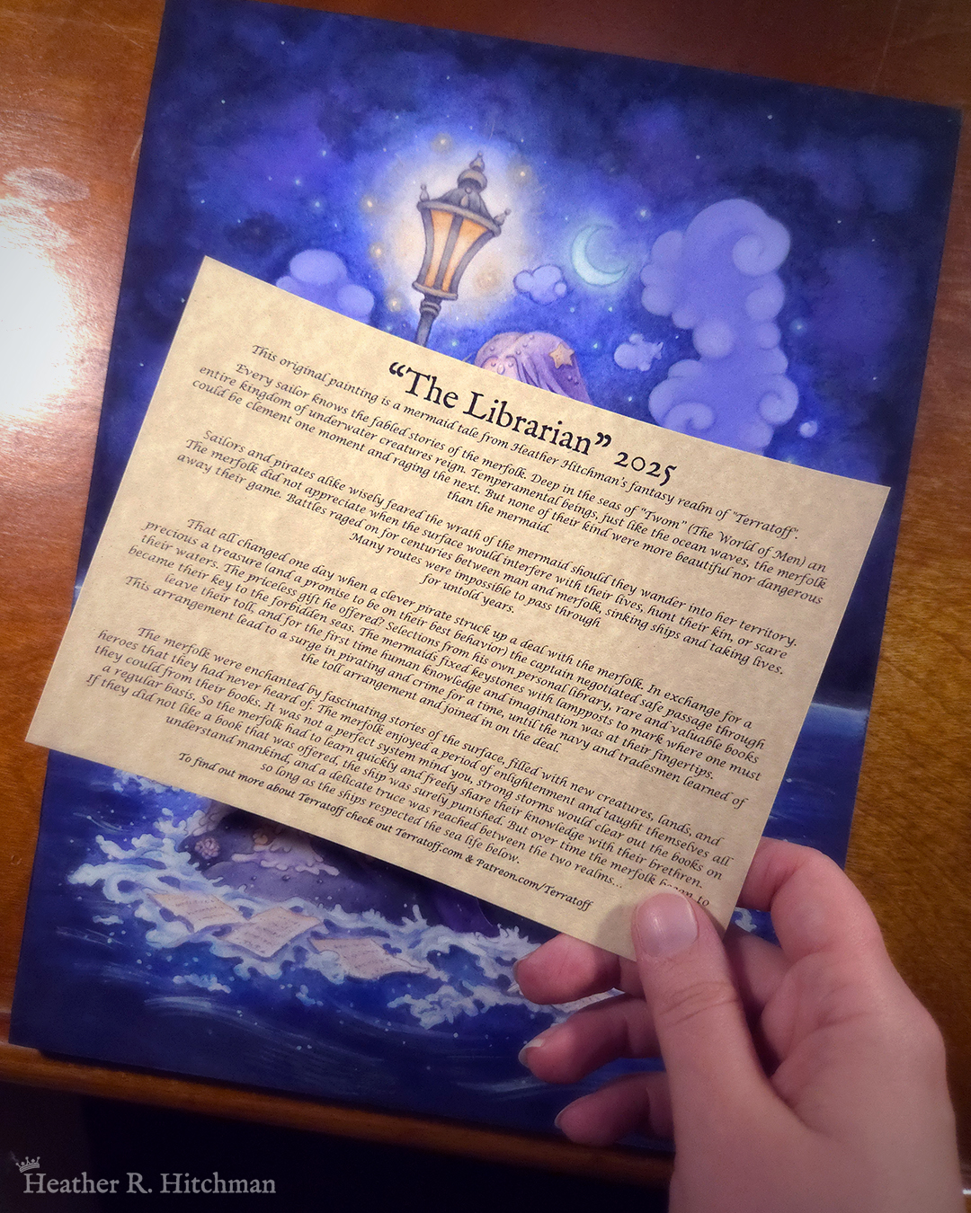 Photograph of the artist Heather R. Hitchman’s hand holding up a parchment page over her original painting “The Librarian”. The parchment covers most of the painting, leaving only the blue sky, waves, and a glowing lantern visible. The page has the story of “The Librarian” printed on it in black ink.
