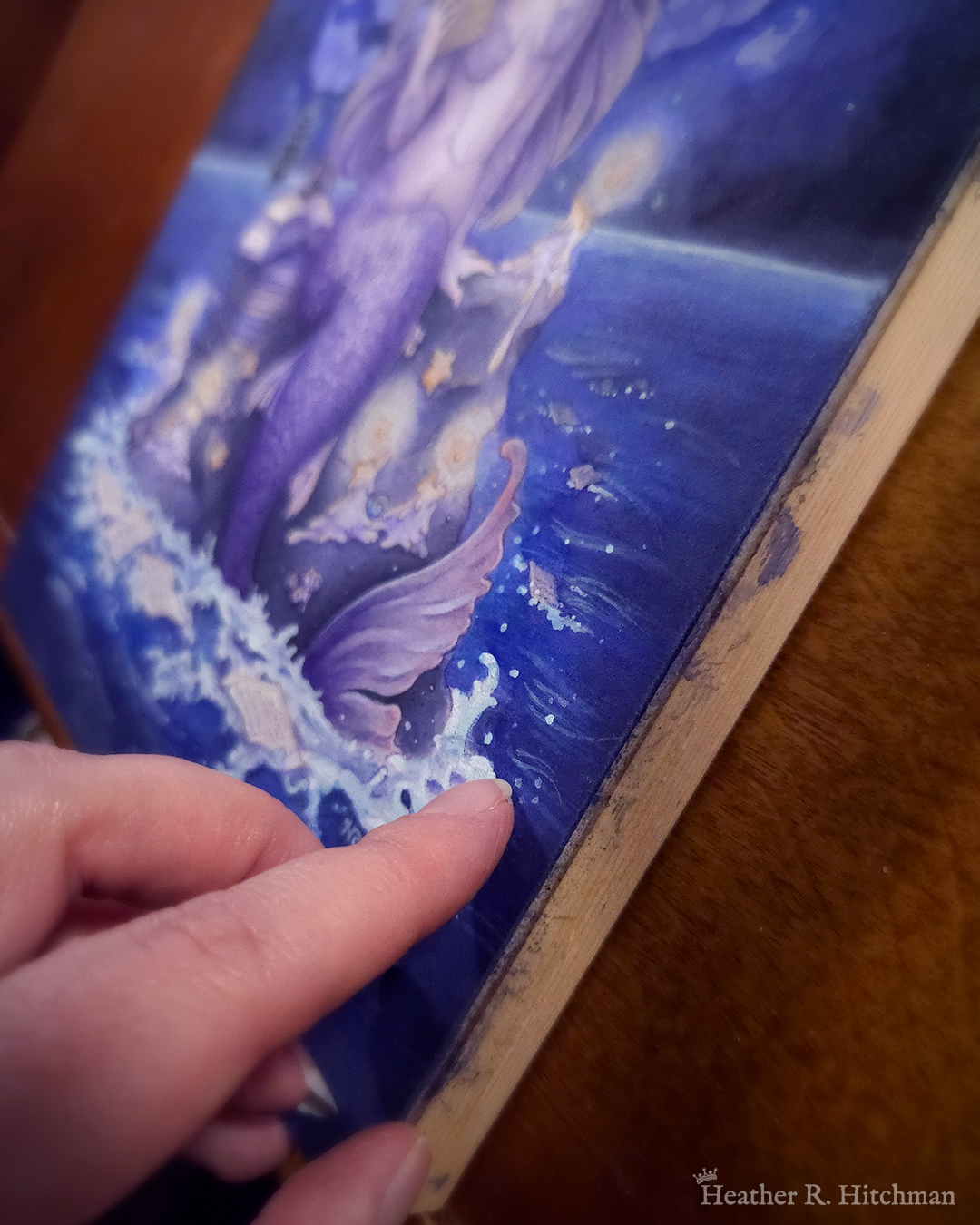 Photograph of the side view of the artist Heather R. Hitchman’s “The Librarian”. This viewpoint shows the 1 inch thickness of the birch board that the painting is mounted on. The photograph shows part of the painting, the tail of a purple mermaid sitting on a rock in the middle of a blue sea is visible.