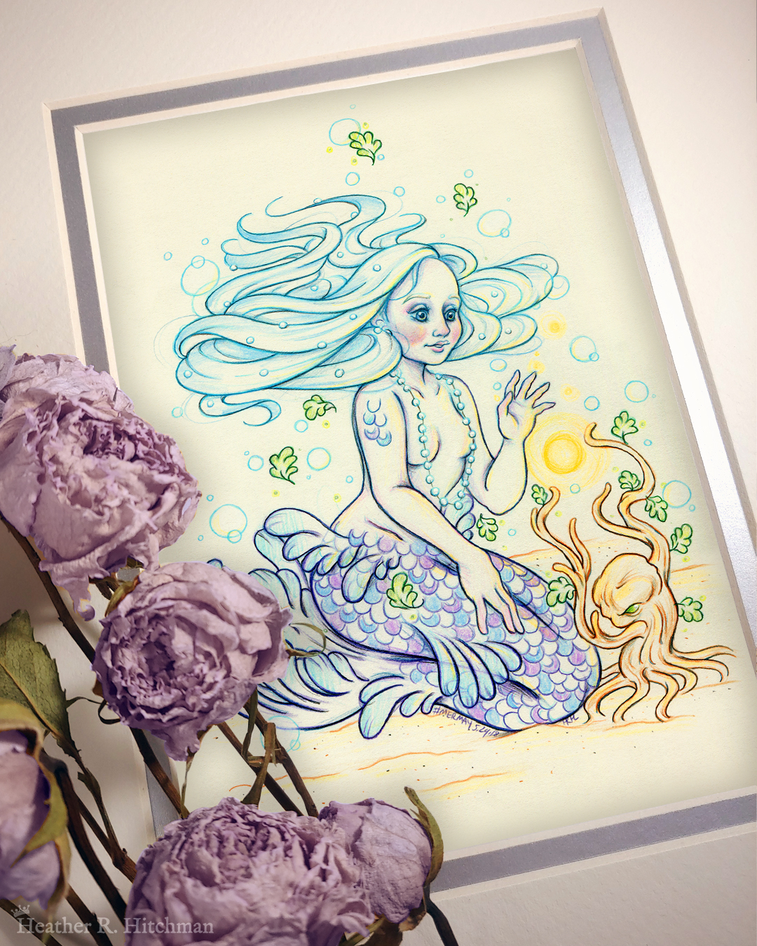Photograph of a matted print of a colored pencil and graphite full body drawing of a purple and blue mermaid sitting on top of her tail in the sand at the bottom of the sea. The mermaid has light purple and blue scales on her tail and shoulders, and flowing light blue hair that is hovering around her head. She sits at a ¾ position facing to the right. A string of white pearls are worn around the mermaid’s neck that discreetly cover her bare chest. Gazing with a mystified expression towards a strange woody plant growing in the sand, she reaches forward towards a round orb of golden light hovering between herself and the plant. The mat-board is white on the top layer and silver on the second layer. Purple roses rest on top of the print to the left. A small silver stamp with the Terratoff icon is at the bottom left corner of the print, under the roses.