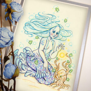 Photograph of a matted print of a colored pencil and graphite full body drawing of a purple and blue mermaid sitting on top of her tail in the sand at the bottom of the sea. The mermaid has light purple and blue scales on her tail and shoulders, and flowing light blue hair that is hovering around her head. She sits at a ¾ position facing to the right. A string of white pearls are worn around the mermaid’s neck that discreetly cover her bare chest. Gazing with a mystified expression towards a strange woody plant growing in the sand, she reaches forward towards a round orb of golden light hovering between herself and the plant. The mat-board is white on the top layer and silver on the second layer. Blue roses rest on top of the print to the left. A small silver stamp with the Terratoff icon is at the bottom left corner of the print, under the roses.