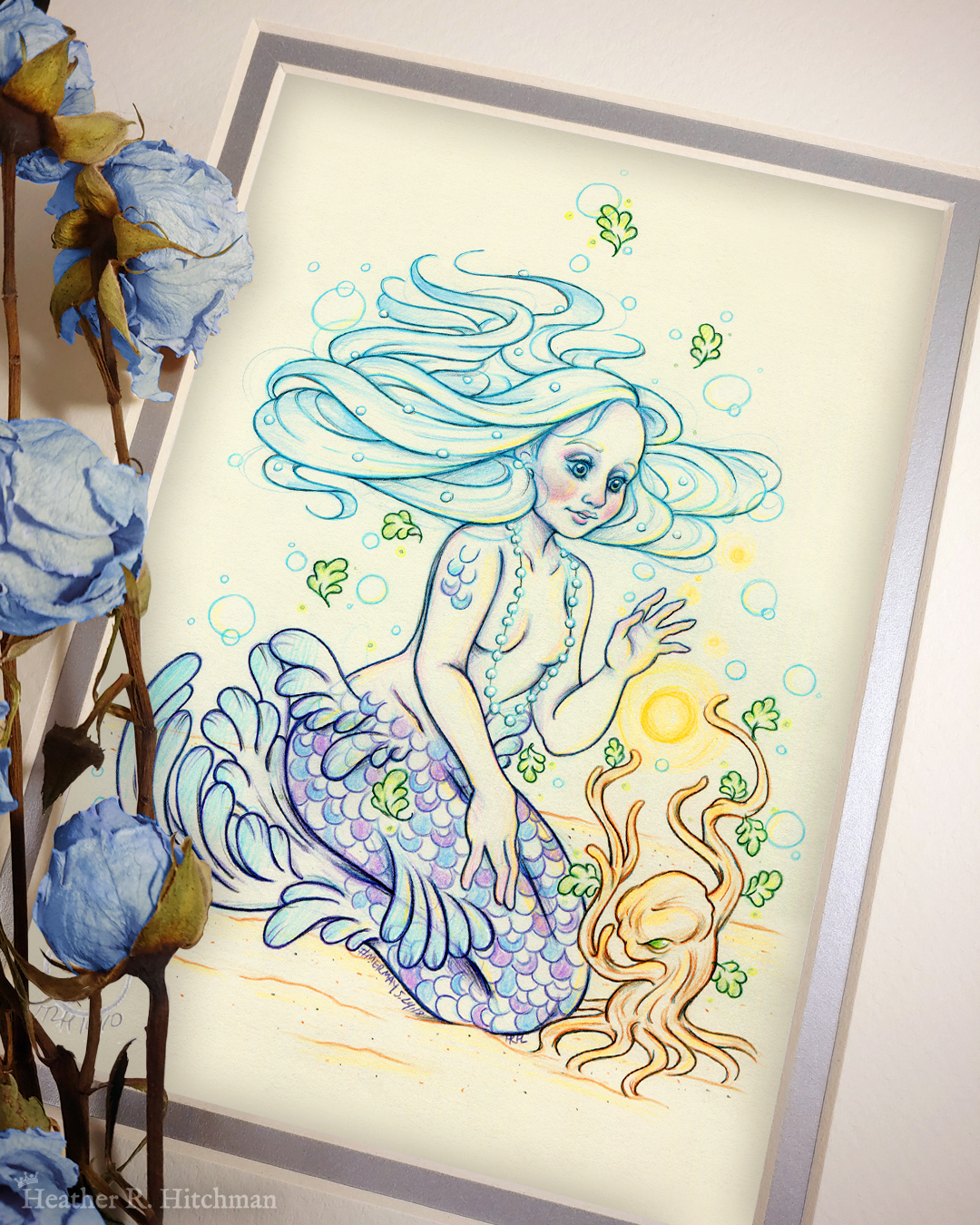 Photograph of a matted print of a colored pencil and graphite full body drawing of a purple and blue mermaid sitting on top of her tail in the sand at the bottom of the sea. The mermaid has light purple and blue scales on her tail and shoulders, and flowing light blue hair that is hovering around her head. She sits at a ¾ position facing to the right. A string of white pearls are worn around the mermaid’s neck that discreetly cover her bare chest. Gazing with a mystified expression towards a strange woody plant growing in the sand, she reaches forward towards a round orb of golden light hovering between herself and the plant. The mat-board is white on the top layer and silver on the second layer. Blue roses rest on top of the print to the left. A small silver stamp with the Terratoff icon is at the bottom left corner of the print, under the roses.