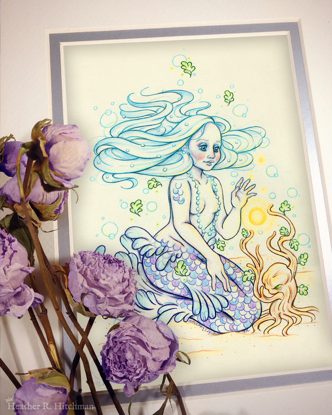 Photograph of a matted print of a colored pencil and graphite full body drawing of a purple and blue mermaid sitting on top of her tail in the sand at the bottom of the sea. The mermaid has light purple and blue scales on her tail and shoulders, and flowing light blue hair that is hovering around her head. She sits at a ¾ position facing to the right. A string of white pearls are worn around the mermaid’s neck that discreetly cover her bare chest. Gazing with a mystified expression towards a strange woody plant growing in the sand, she reaches forward towards a round orb of golden light hovering between herself and the plant. The mat-board is white on the top layer and silver on the second layer. Purple roses rest on top of the print to the left. A small silver stamp with the Terratoff icon is at the bottom left corner of the print, under the roses.