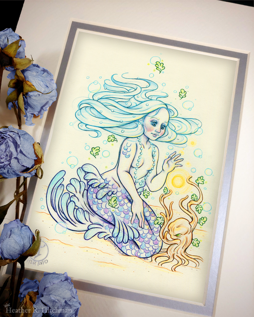 Photograph of a matted print of a colored pencil and graphite full body drawing of a purple and blue mermaid sitting on top of her tail in the sand at the bottom of the sea. The mermaid has light purple and blue scales on her tail and shoulders, and flowing light blue hair that is hovering around her head. She sits at a ¾ position facing to the right. A string of white pearls are worn around the mermaid’s neck that discreetly cover her bare chest. Gazing with a mystified expression towards a strange woody plant growing in the sand, she reaches forward towards a round orb of golden light hovering between herself and the plant. The mat-board is white on the top layer and silver on the second layer. Blue roses rest on top of the print to the left. A small silver stamp with the Terratoff icon is at the bottom left corner of the print, under the roses.