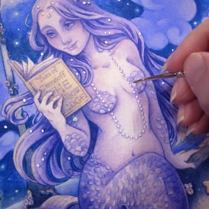 Close up photograph of a print of a watercolor and gouache full body painting of a purple mermaid reading a book under the moonlight. In the background puffy light blue fish shaped clouds fill the night sky surrounding a crescent moon. The mermaid is holding a book in her right hand up to her face. Her other hand is placed behind her as she leans back slightly on a gray rock, her left arm supporting her weight. Her body faces the viewer, and she has a peaceful smile as she looks towards the book and reads. The print has been embellished with details of silver paint on some of the mermaid’s scales, the pearls in her hair and around her neck. The fingertips of the artist are in the right section of the photograph using a small paintbrush to paint silver pigment onto the mermaid’s scales on her chest.