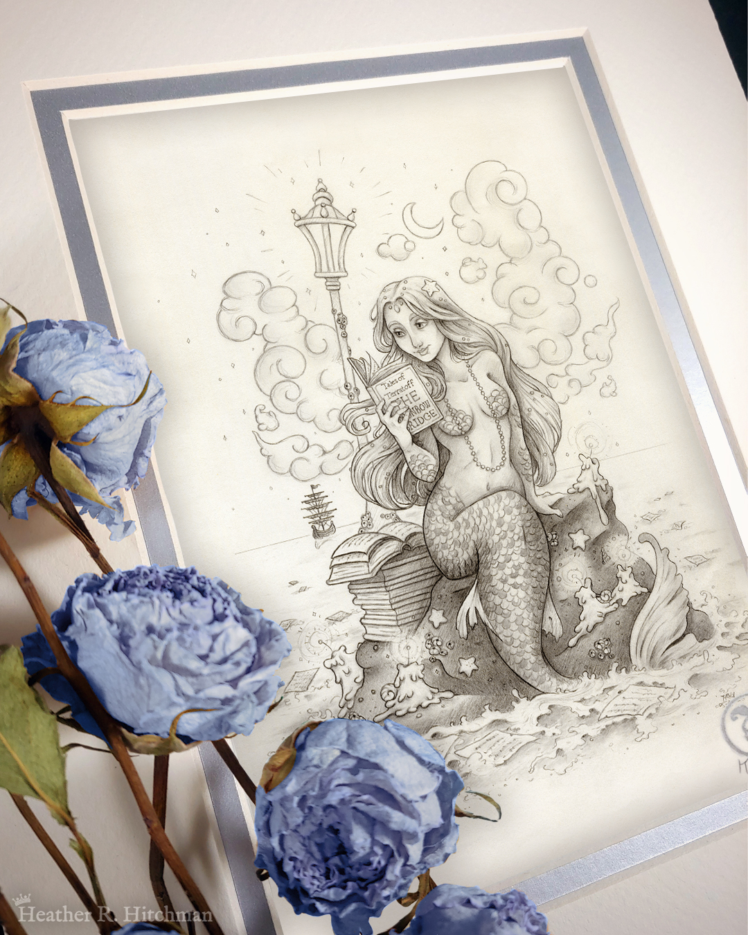 Photograph of a matted print of a black and white graphite full body drawing of a mermaid reading a book under the moonlight while sitting on a rock in the middle of the sea. The rock is covered in glowing candles and also has a pile of books besides the mermaid’s tail. An old fashioned street lantern covered in barnacles is attached to the rock, illuminating the night around the mermaid’s face. Gentle waves wash up against the rock. Loose pages from books float in the water into the distance, leading towards a mysterious ship that vanishes into the night. Puffy fish shaped clouds fill the nigh sky surrounding a crescent moon. The mermaid is holding a book in her right hand up to her face. Her other hand is placed behind her as she leans back slightly, her left arm supporting her weight. Her body faces the viewer, and she has a peaceful smile as she looks towards the book and reads. Blue roses rest on top of the print to the left. A small silver stamp with the Terratoff icon is at the bottom right corner of the print.