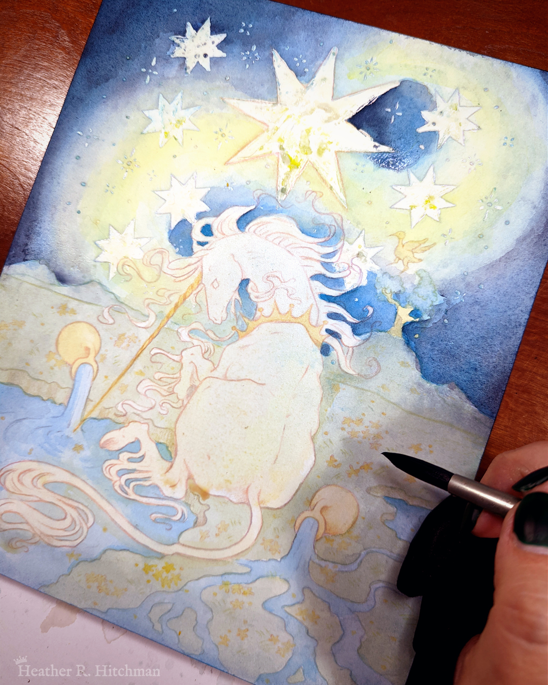 Process photo of a loose watercolor and gouache painting of a unicorn sitting by a lake under a night sky. The sky is dark blue and filled with tiny white stars. Seven large white stars surround a massive yellow star in the center of the sky. A swirl of bright yellow light weaves upwards towards the heavens, illuminating the scene below. The unicorn sits in front of a landscape scene, with green mountaintops in the background. On one of the mountaintops a small tree grows, on top of the branches a yellow ibis bird flaps its wings. The unicorn is sitting with its back towards the viewer, the head turned in profile as she dips her long golden horn into the lake to the left of the image. The unicorn wears a golden crown around her neck. Two golden urns rest on their sides on the ground, spilling water onto the grassy scene that trickles into the lake waters. The artist's hand is painting in the bottom right corner.
