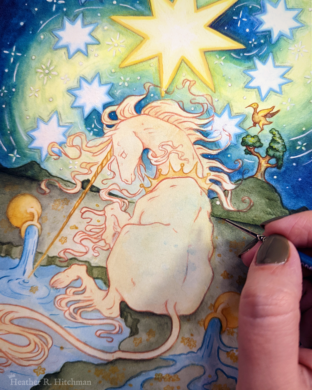 Process photo of a loose watercolor and gouache painting of a unicorn sitting by a lake under a night sky. The sky is dark blue and filled with tiny white stars. Seven large white stars surround a massive yellow star in the center of the sky. A swirl of bright yellow light weaves upwards towards the heavens, illuminating the scene below. The unicorn sits in front of a landscape scene, with green mountaintops in the background. On one of the mountaintops a small tree grows, on top of the branches a yellow ibis bird flaps its wings. The unicorn is sitting with its back towards the viewer, the head turned in profile as she dips her long golden horn into the lake to the left of the image. The unicorn wears a golden crown around her neck. Two golden urns rest on their sides on the ground, spilling water onto the grassy scene that trickles into the lake waters. The artist's hand is painting in the bottom right corner.