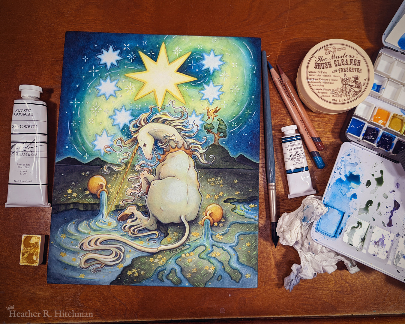 Photograph of a watercolor and gouache painting of a unicorn sitting by a lake under a night sky. The sky is dark blue and filled with tiny white stars. Seven large white stars surround a massive yellow star in the center of the sky. A swirl of bright yellow light weaves upwards towards the heavens, illuminating the scene below. The unicorn sits in front of a landscape scene, with green mountaintops in the background. On one of the mountaintops a small tree grows, on top of the branches a yellow ibis bird flaps its wings. The unicorn is sitting with its back towards the viewer, the head turned in profile as she dips her long golden horn into the lake to the left of the image. The unicorn wears a golden crown around her neck. Two golden urns rest on their sides on the ground, spilling water onto the grassy scene that trickles into the lake waters. The painting is resting on a wooden tabletop, with watercolor brushes and supplies surrounding it.