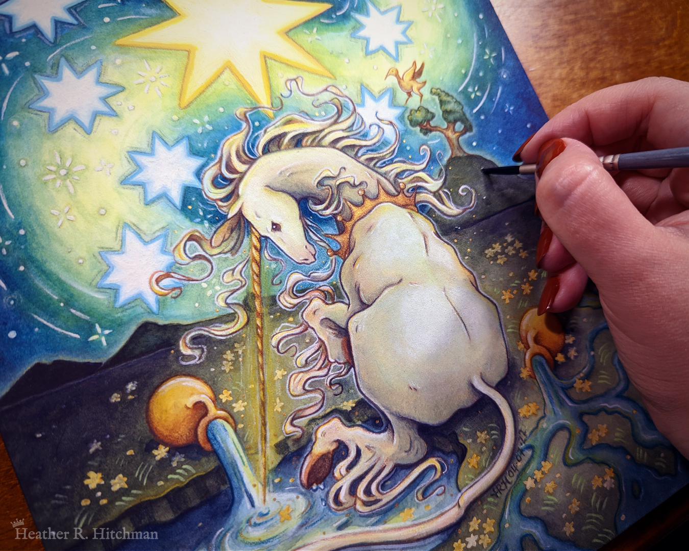 Photo of the artist's hand painting a watercolor and gouache painting of a unicorn sitting by a lake under a night sky. Part of the hand is visible to the right, and holds a paintbrush over the painting. The sky is dark blue and filled with tiny white stars. Seven large white stars surround a massive yellow star in the center of the sky. A swirl of bright yellow light weaves upwards towards the heavens, illuminating the scene below. The unicorn sits in front of a landscape scene, with green mountaintops in the background. On one of the mountaintops a small tree grows, on top of the branches a yellow ibis bird flaps its wings. The unicorn is sitting with its back towards the viewer, the head turned in profile as she dips her long golden horn into the lake to the left of the image. The unicorn wears a golden crown around her neck. Two golden urns rest on their sides on the ground, spilling water onto the grassy scene that trickles into the lake waters.