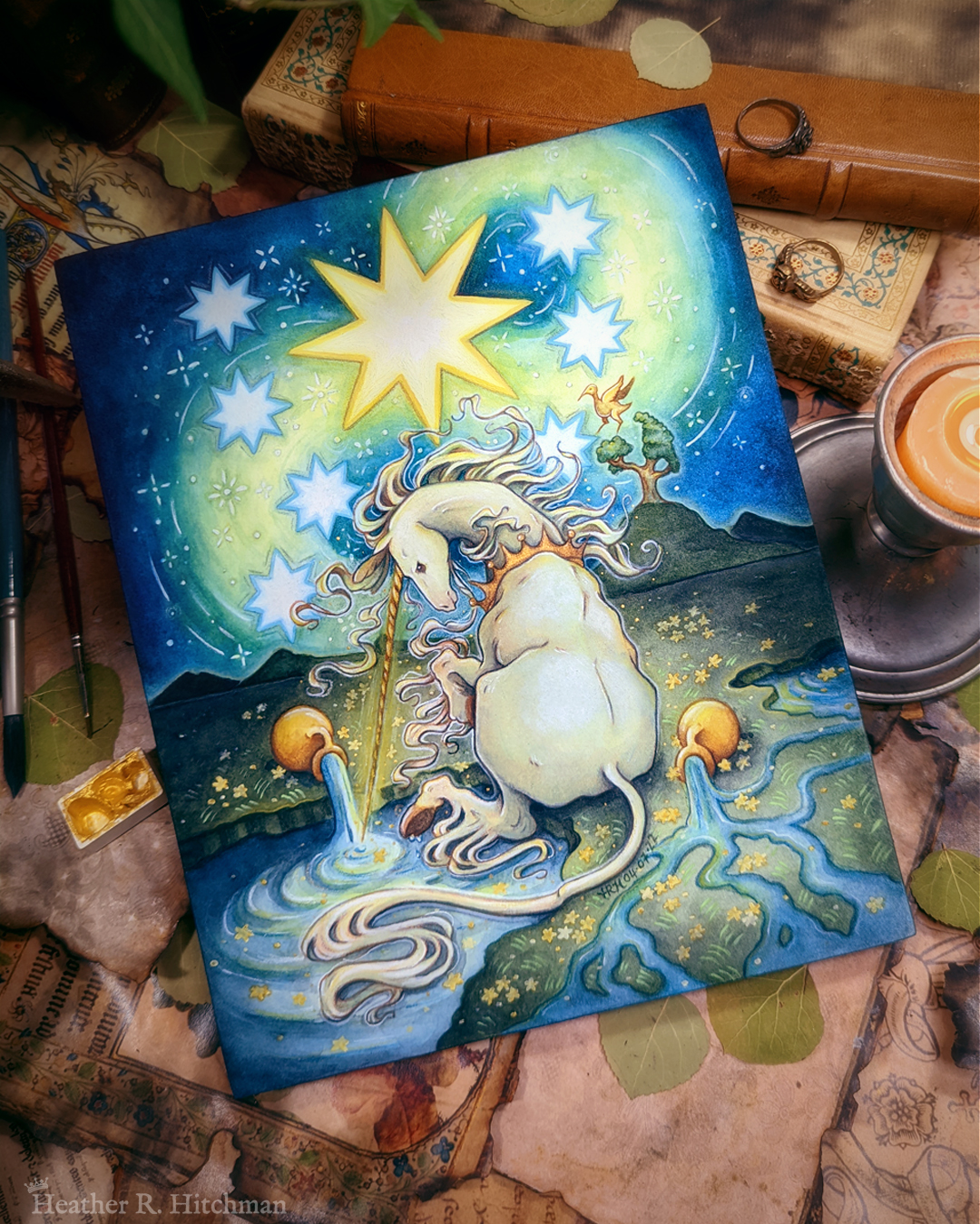 Watercolor and gouache painting of a unicorn sitting by a lake under a night sky. The sky is dark blue and filled with tiny white stars. Seven large white stars surround a massive yellow star in the center of the sky. A swirl of bright yellow light weaves upwards towards the heavens, illuminating the scene below. The unicorn sits in front of a landscape scene, with green mountaintops in the background. On one of the mountaintops a small tree grows, on top of the branches a yellow ibis bird flaps its wings. The unicorn is sitting with its back towards the viewer, the head turned in profile as she dips her long golden horn into the lake to the left of the image. The unicorn wears a golden crown around her neck. Two golden urns rest on their sides on the ground, spilling water onto the grassy scene that trickles into the lake waters. The painting is placed on a table covered with book pages and candles.