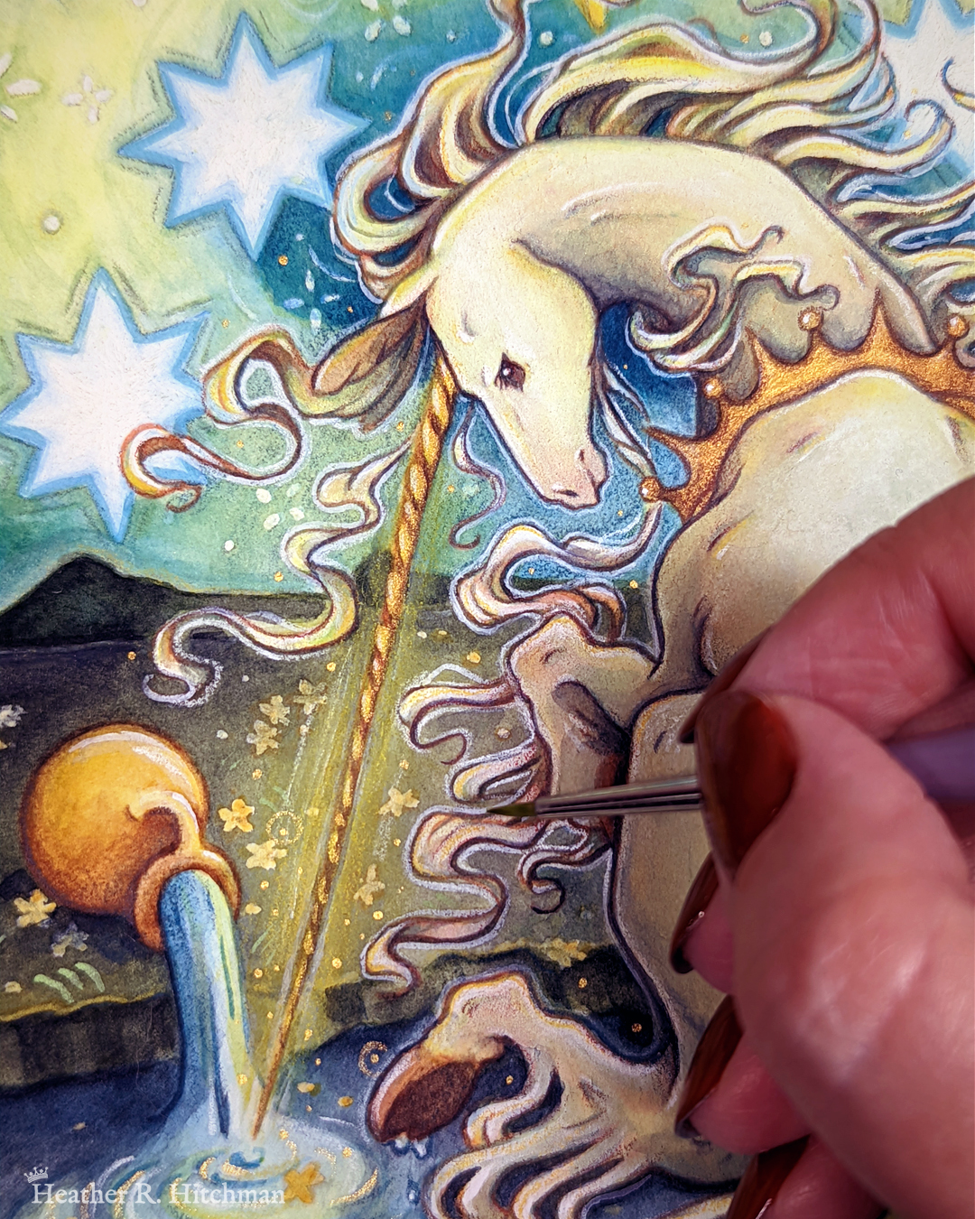 Close up process photo of a loose watercolor and gouache painting of a unicorn sitting by a lake under a night sky. The sky is dark blue and filled with tiny white stars. Several large white stars are visible. A swirl of bright yellow light weaves upwards towards the heavens, illuminating the scene below. The unicorn sits in front of a landscape scene, with green mountaintops in the background. The unicorn is sitting with its back towards the viewer, the head turned in profile as she dips her long golden horn into the lake to the left of the image. The unicorn wears a golden crown around her neck. A golden urn rests on its side on the ground, spilling water onto the grassy scene that trickles into the lake waters. The artist’s hand is painting with a paintbrush over the painting in the bottom right.