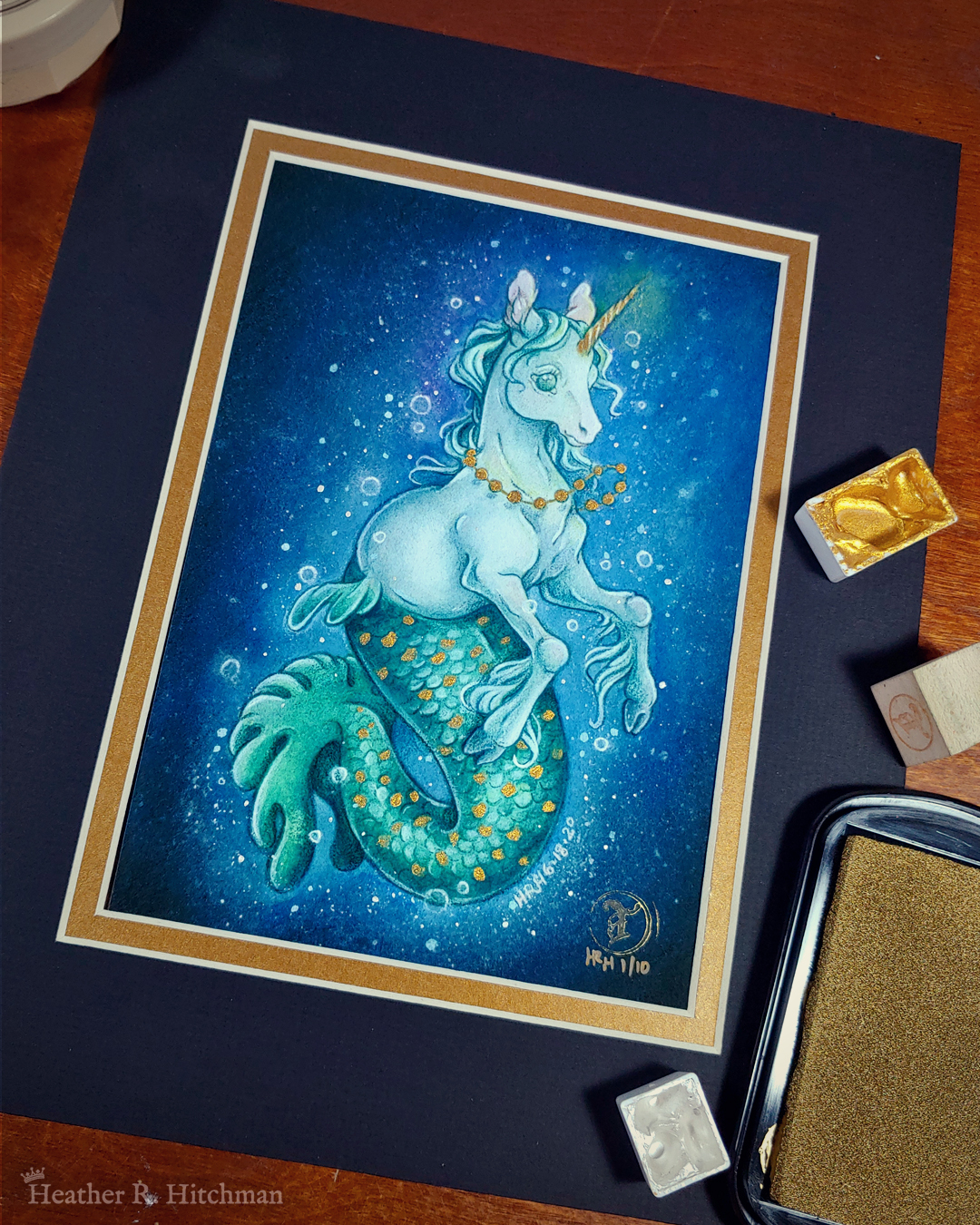 Photograph of a matted print of a full body watercolor, gouache, and colored pencil painting of a mermaid unicorn under the sea. Also known as a mermacorn, the creature has a white unicorn torso and a green mermaid tail. The mermacorn appears to be a young colt, with large light green eyes, a short glowing golden horn, and long spindly front legs. The unicorn wears a necklace of golden pearls and floats effortlessly in the dark waters with a smile on its face. They are facing ¾ to the right, and appear to be glowing so that the dark waters immediately around them are illuminated to a lighter blue color. The mermacorn has golden scales scattered among the length of its green tail. The background is a gradient of blue water, with white bubbles surrounding the floating mermacorn from head to tail. The mat-board is black on the top layer and gold on the second layer. Small pans of gold and silver watercolor pigment and a golden ink pad surround the print.