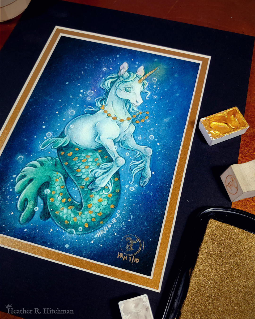 Photograph of a matted print of a full body watercolor, gouache, and colored pencil painting of a mermaid unicorn under the sea. Also known as a mermacorn, the creature has a white unicorn torso and a green mermaid tail. The mermacorn appears to be a young colt, with large light green eyes, a short glowing golden horn, and long spindly front legs. The unicorn wears a necklace of golden pearls and floats effortlessly in the dark waters with a smile on its face. They are facing ¾ to the right, and appear to be glowing so that the dark waters immediately around them are illuminated to a lighter blue color. The mermacorn has golden scales scattered among the length of its green tail. The background is a gradient of blue water, with white bubbles surrounding the floating mermacorn from head to tail. The mat-board is black on the top layer and gold on the second layer. Small pans of gold and silver watercolor pigment and a golden ink pad surround the print.